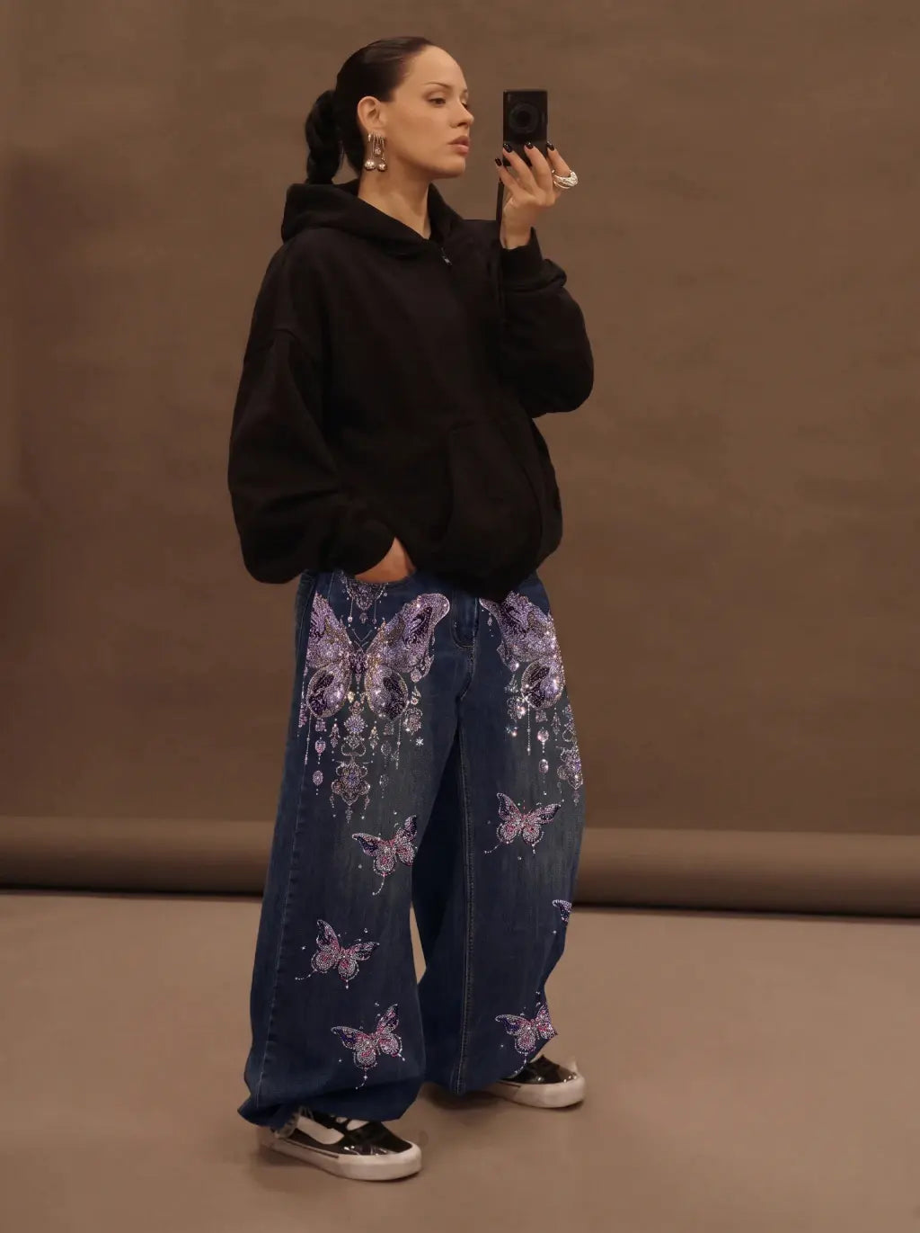 A woman takes a mirror selfie wearing a black oversized hoodie and wide-leg blue jeans with purple butterfly and floral prints, paired with black and white sneakers, against a plain brown studio backdrop. TCH Rhinestone Butterfly Charm Wide-Leg Denim Jeans