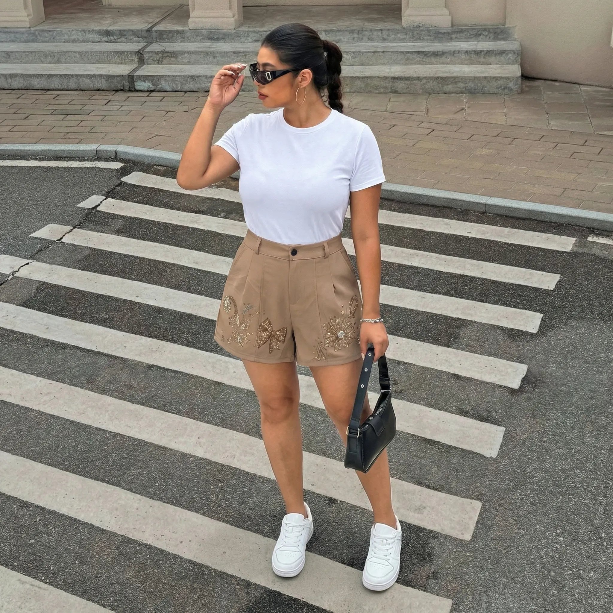 A woman stands on a city crosswalk wearing a white short-sleeve crewneck t-shirt, beige high-waisted shorts with floral embroidery, white sneakers, and holding a small black handbag. TCH Rhinestone Butterfly Floral Embellished Tailored Shorts