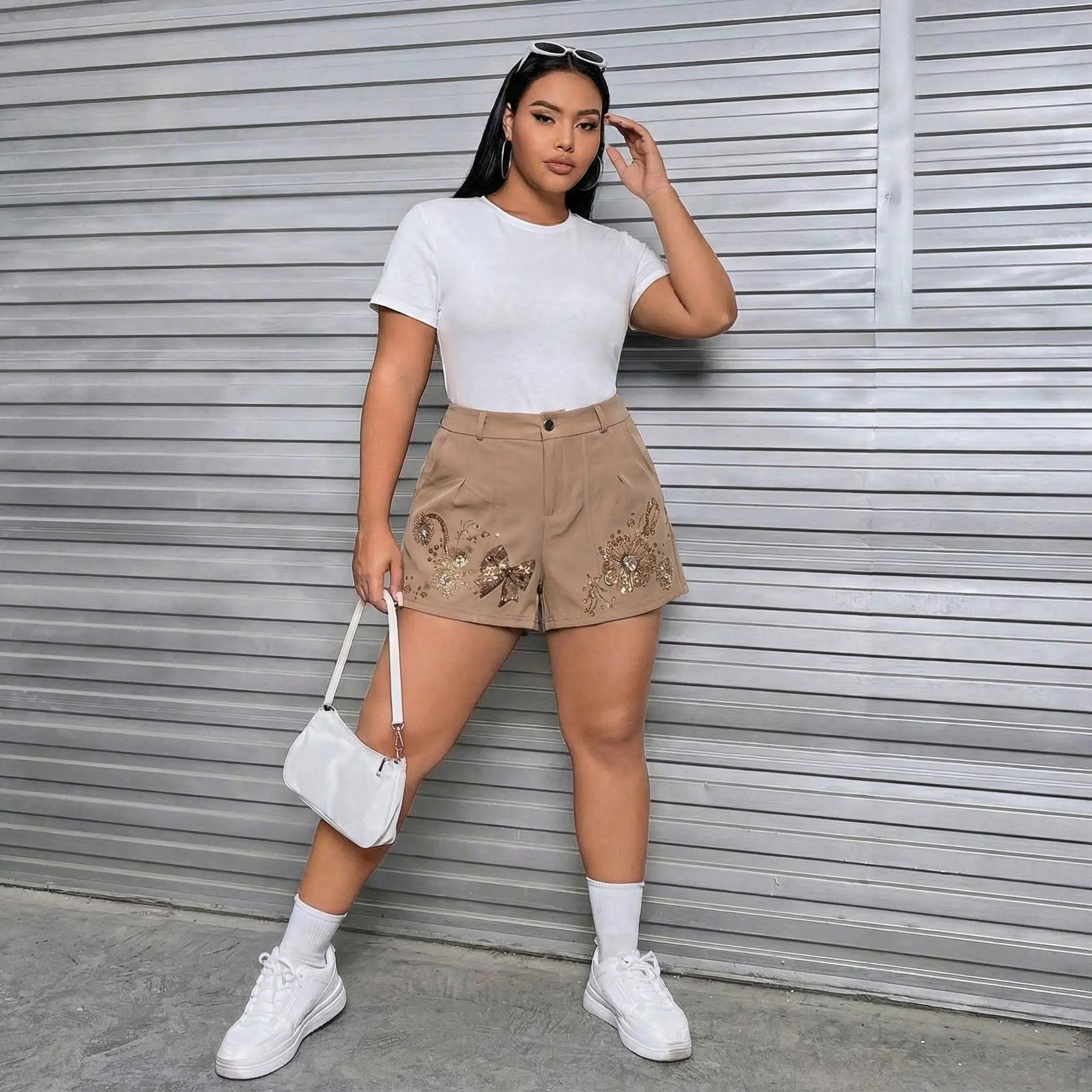 A woman stands in front of a gray corrugated metal wall wearing a white short-sleeve crewneck t-shirt, beige shorts with brown floral embroidery, white socks, white sneakers, and holding a small white handbag. TCH Rhinestone Butterfly Floral Embellished Tailored Shorts