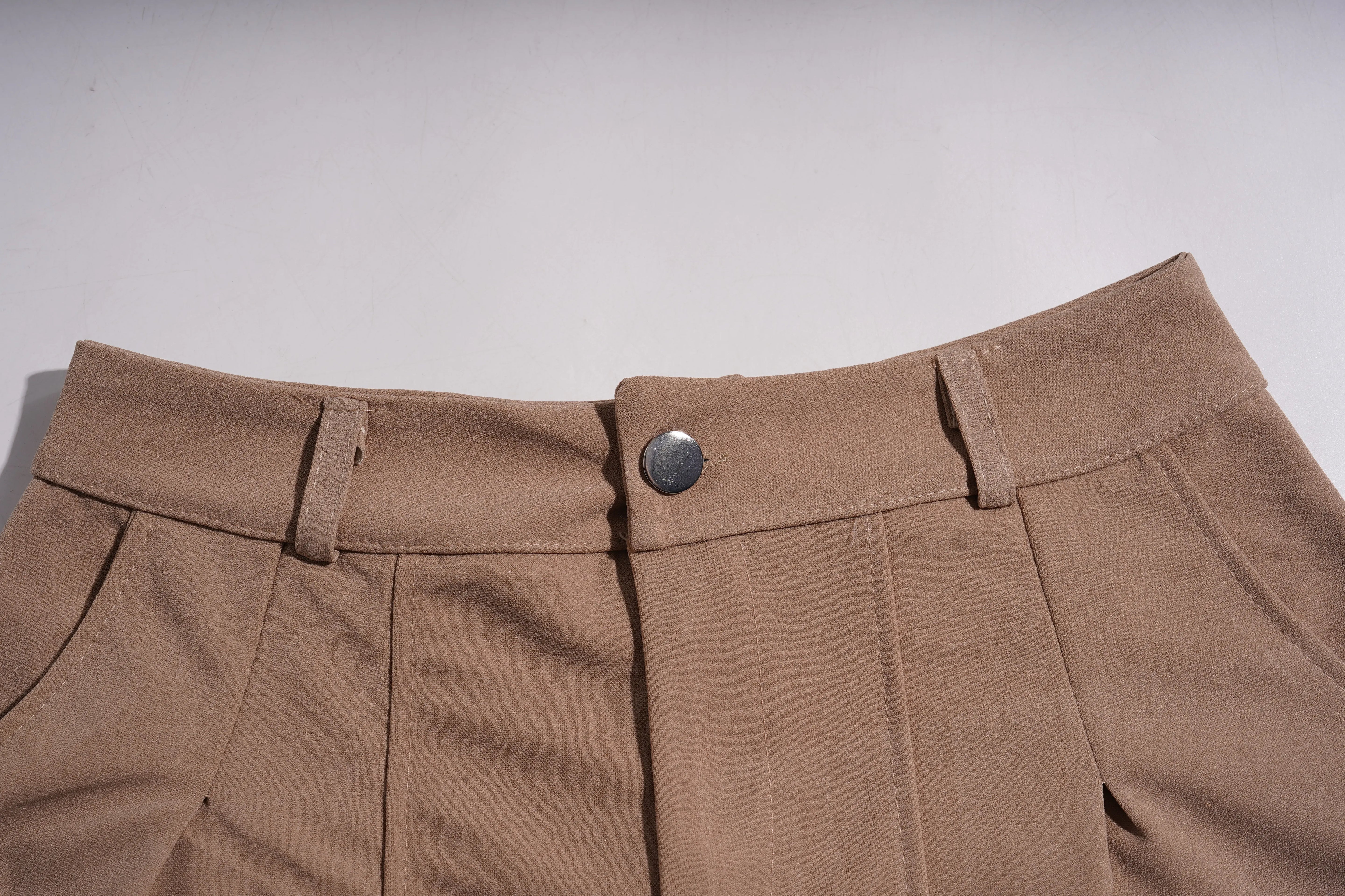 Close-up of the waistband of tan pleated trousers, showing belt loops, a single metal button, and front pleats against a light gray background. TCH Rhinestone Butterfly Floral Embellished Tailored Shorts