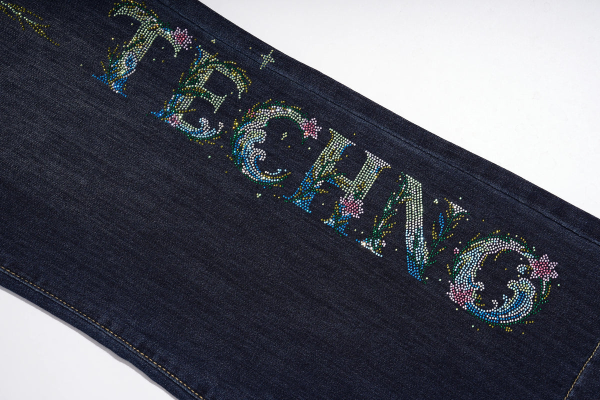 Close-up of a dark blue denim garment featuring the word 'TECHNO' spelled out in large, embellished letters with rhinestones and small pink floral accents, set against a plain white background. TCH Rhinestone Butterfly Graphic Wide-Leg Jeans