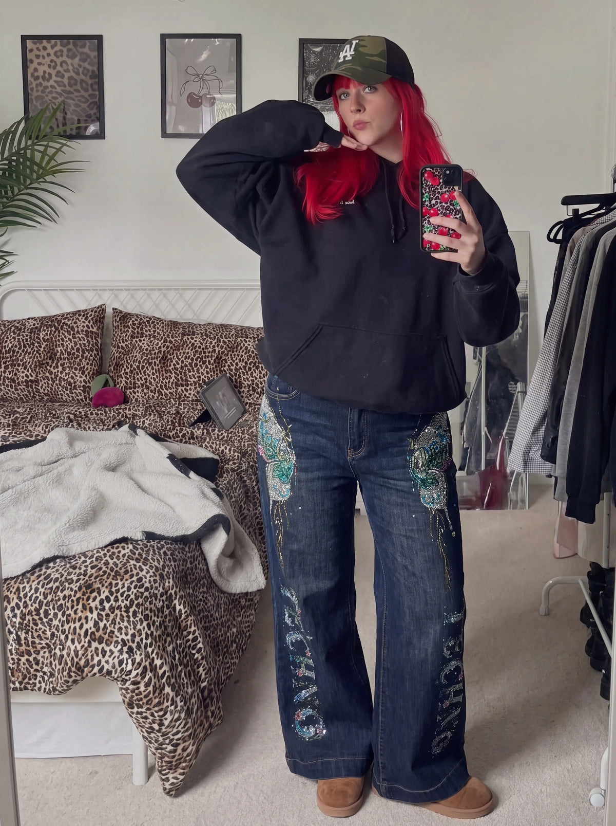A person takes a mirror selfie in a bedroom, wearing a black oversized hoodie, wide-leg blue jeans with colorful embroidered details, a green baseball cap, and brown boots. The room features a bed with leopard-print bedding and a clothing rack in the background. TCH Rhinestone Butterfly Graphic Wide-Leg Jeans
