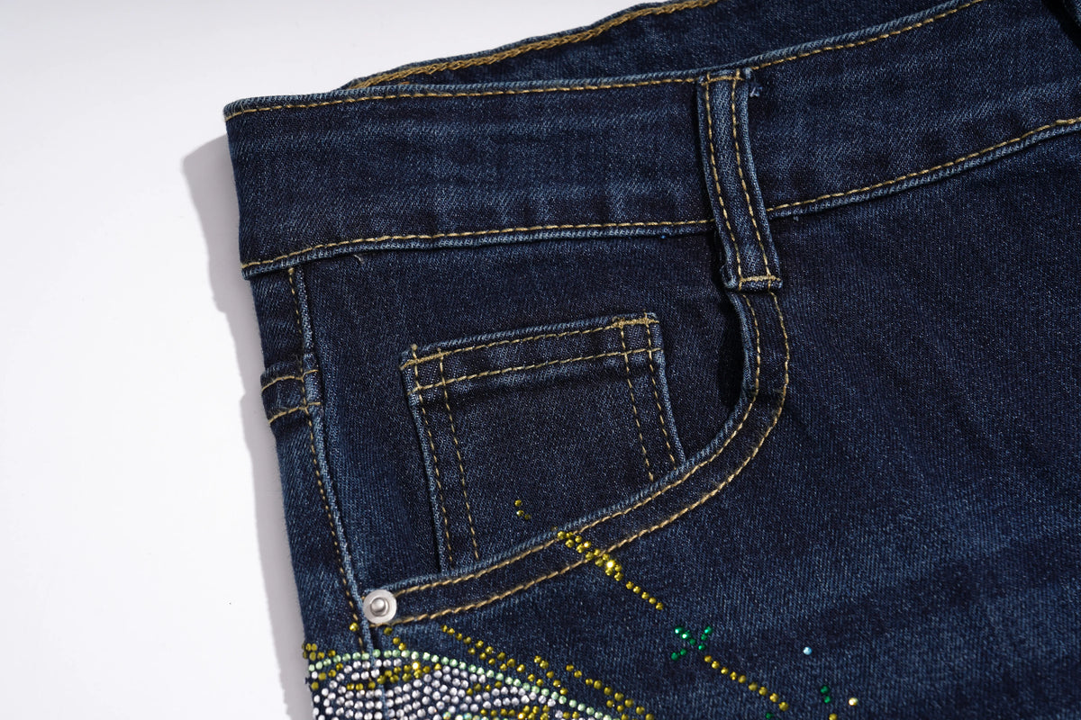 Close-up of the back pocket area of dark blue denim jeans with gold contrast stitching and a decorative embellishment near the bottom corner. TCH Rhinestone Butterfly Graphic Wide-Leg Jeans