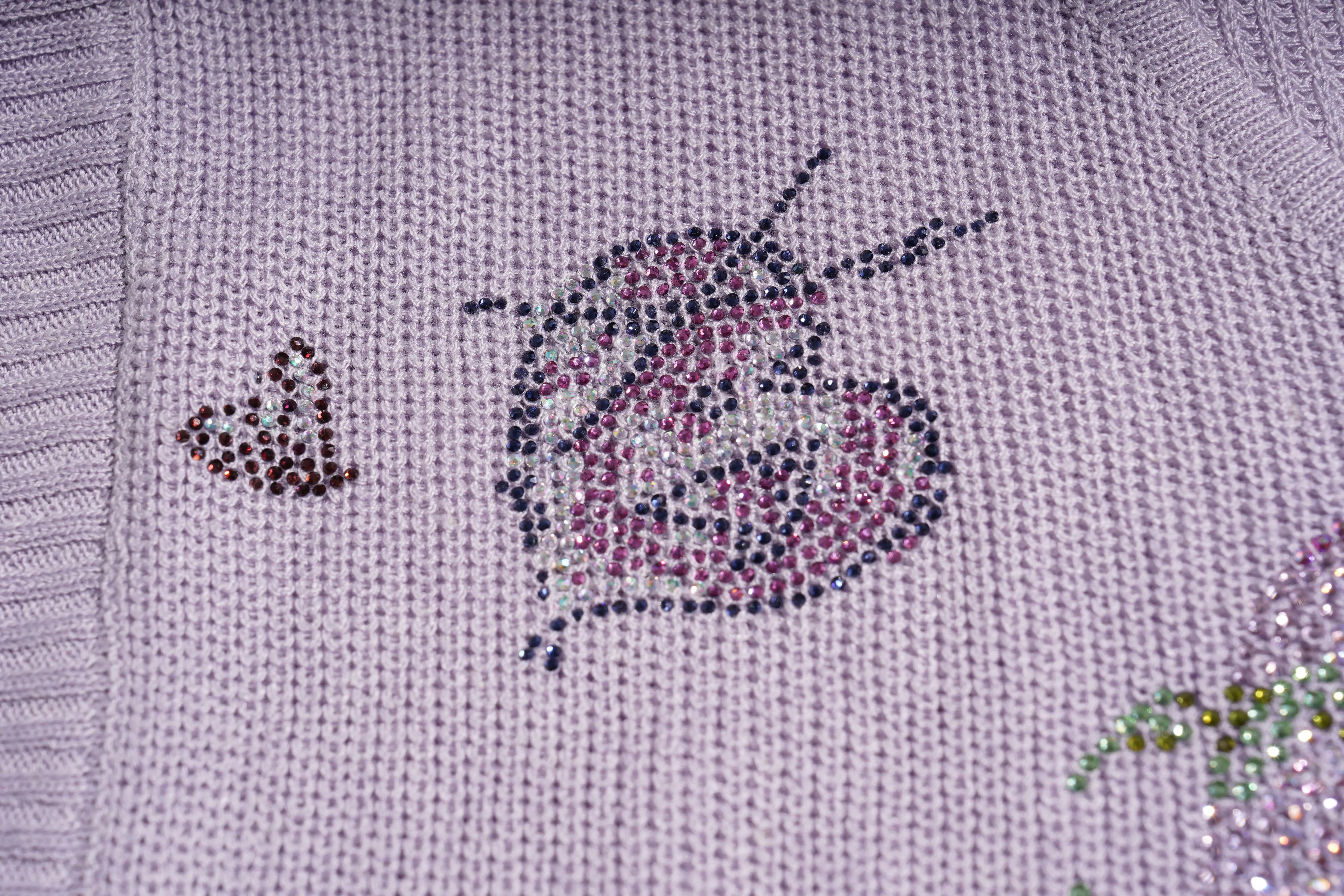 Close-up of a knitted sweater in a pale lavender or light purple color, featuring decorative rhinestone or bead appliques including a butterfly and a butterfly-and-flower motif. TCH Rhinestone Butterfly Knit Twist Top