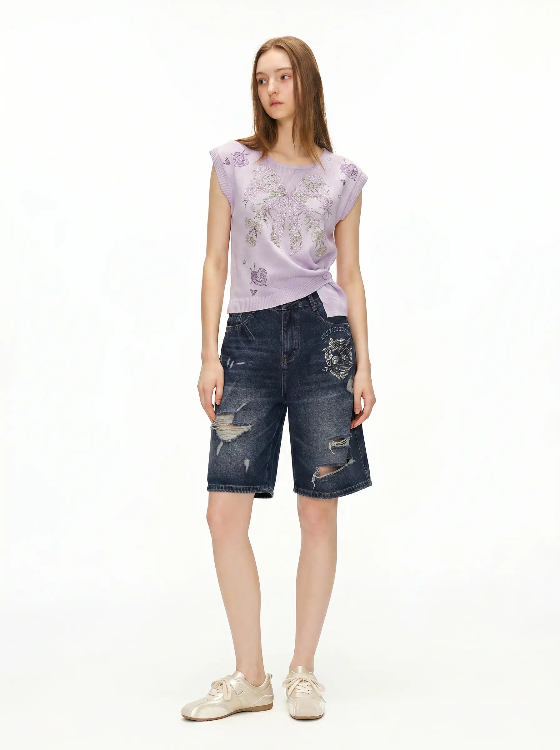 A model stands against a plain white studio background wearing a light lavender sleeveless top with a subtle floral print, dark blue distressed denim shorts, and beige sneakers. TCH Rhinestone Butterfly Knit Twist Top