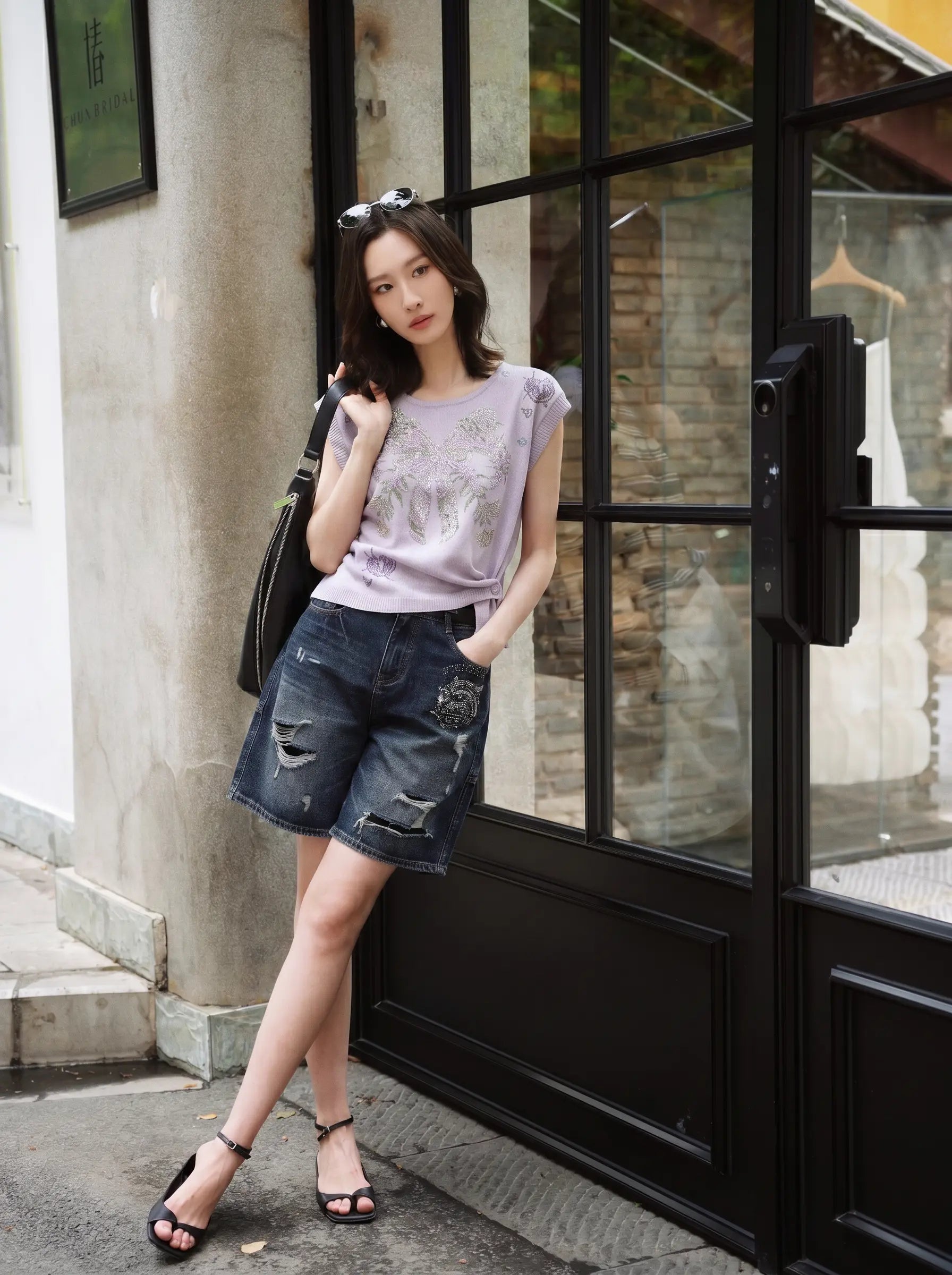 A woman stands outdoors in front of a storefront with black-framed glass doors, wearing a light lavender graphic sleeveless top, distressed dark blue denim shorts, and black strappy sandals, with a black shoulder bag. TCH Rhinestone Butterfly Knit Twist Top