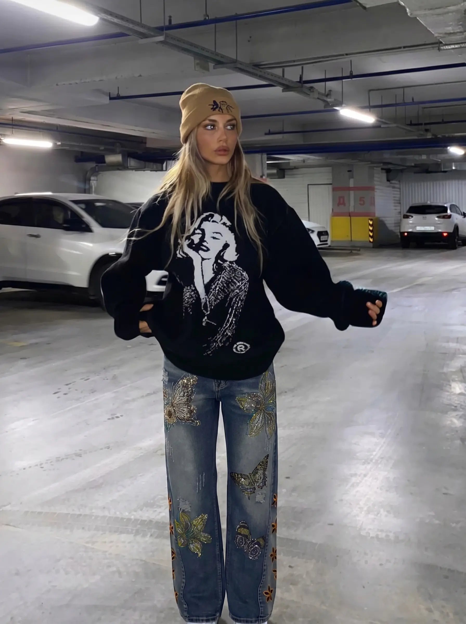 Person wearing a black sweatshirt with a graphic design and floral jeans in a parking garage. TCH Rhinestone Butterfly & Lily Distressed Denim Jeans