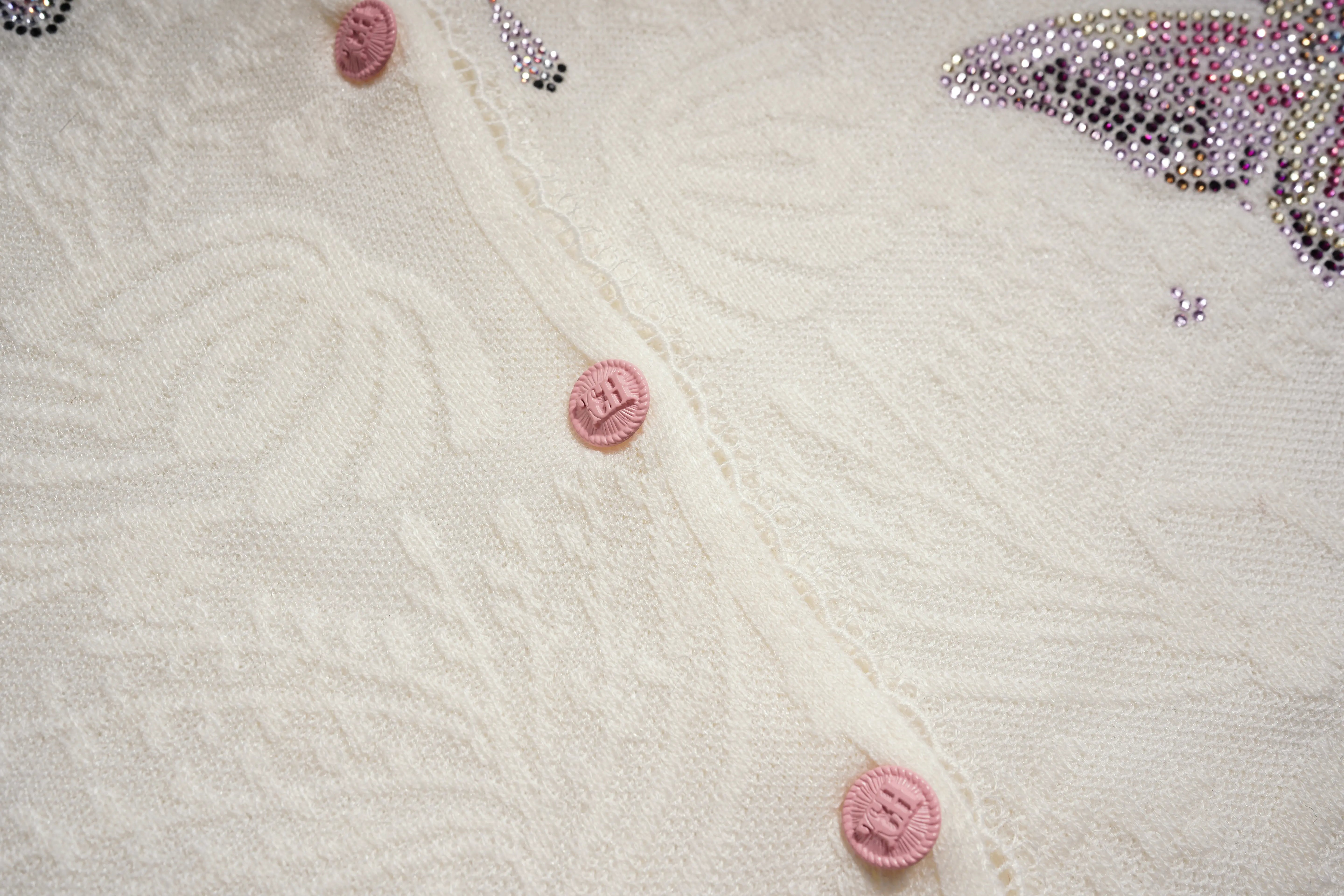 Close-up of a white textured fabric with pink buttons and decorative elements. TCH Rhinestone Butterfly Logo Knit Cardigan