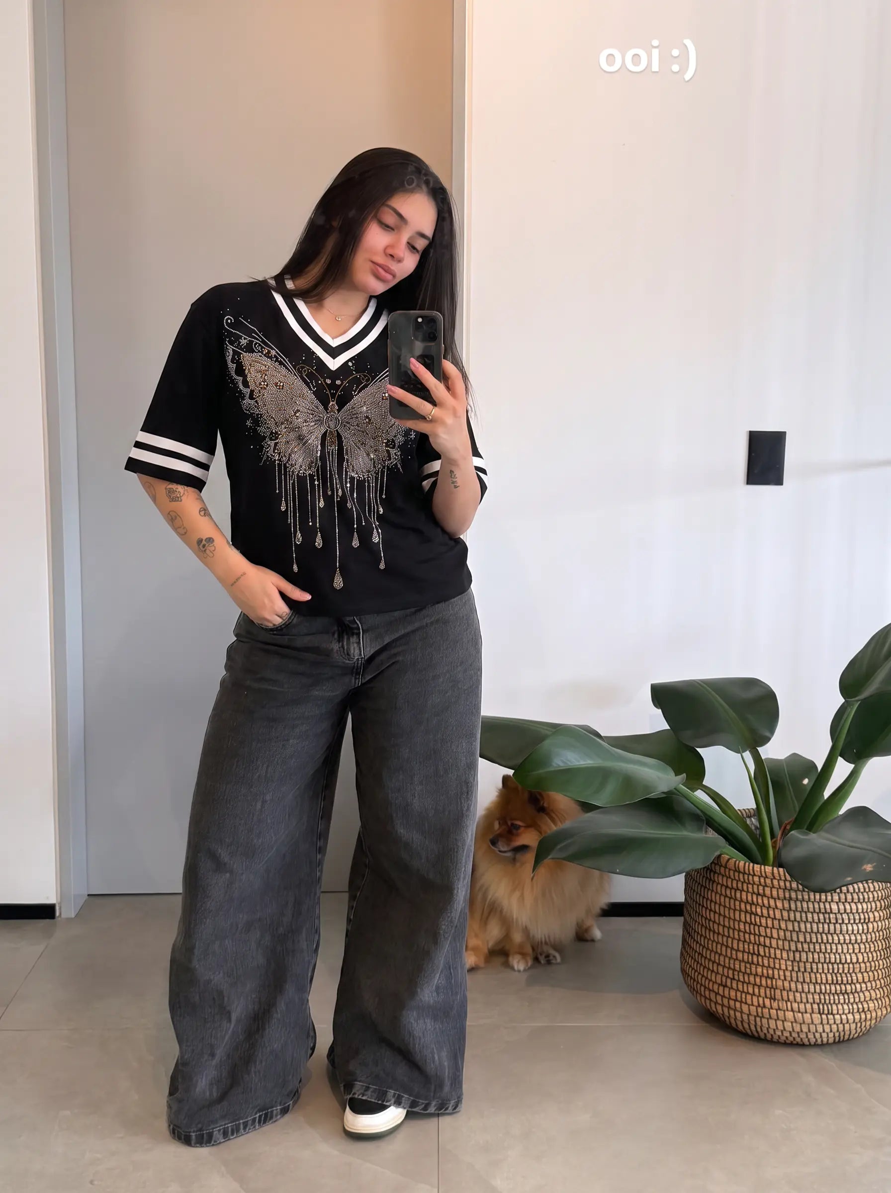 A woman takes a mirror selfie wearing a black V-neck t-shirt with a silver rhinestone butterfly graphic and gray wide-leg jeans. A small dog sits behind her on a tiled floor next to a potted plant. TCH Rhinestone Butterfly V-Neck Oversized Baseball T-Shirt
