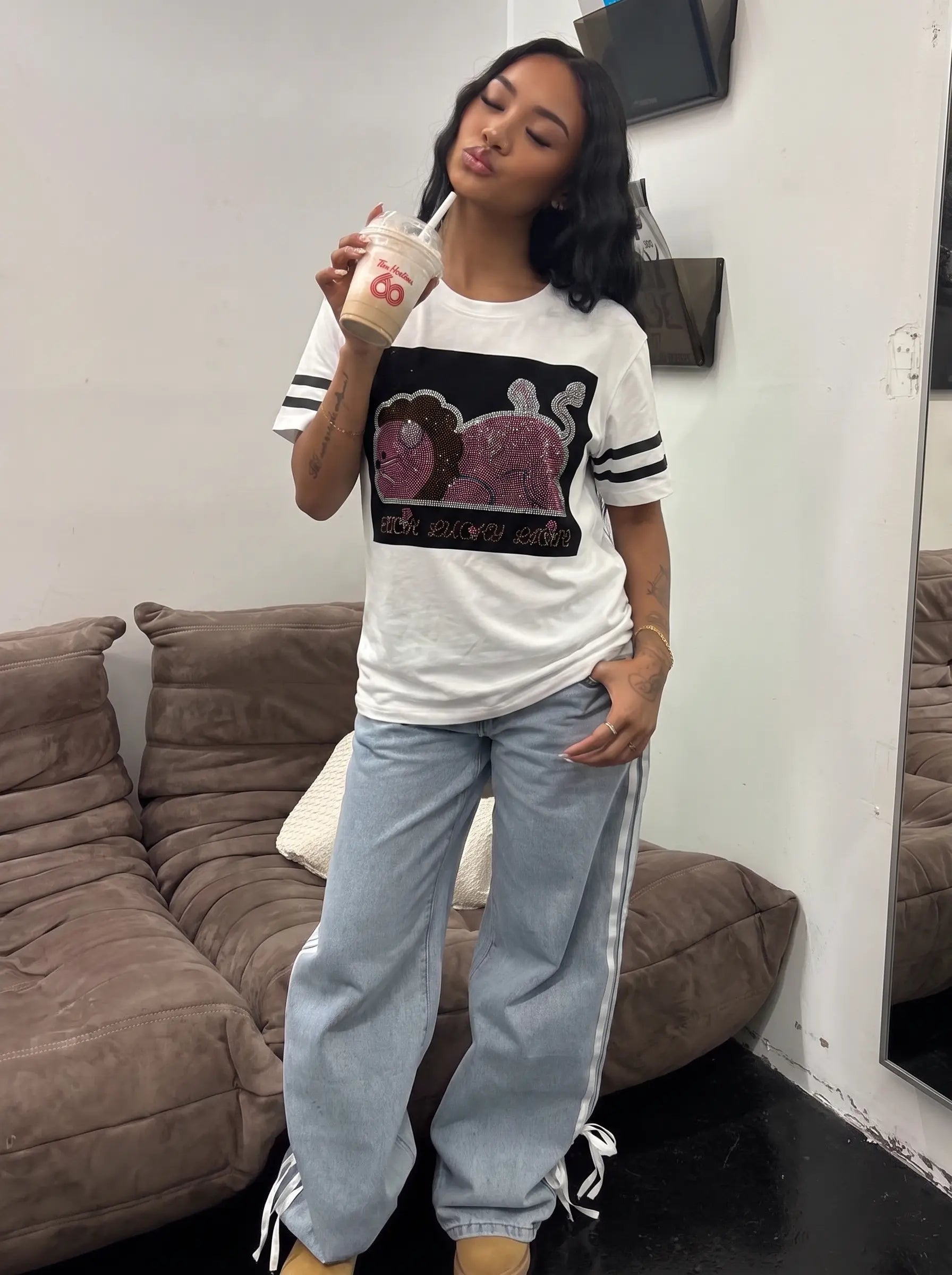 A woman stands indoors wearing a white graphic T-shirt with black and pink cartoon-style artwork and black-and-white striped sleeves, paired with light blue sweatpants with white side stripes and drawstring ankle ties. She holds a takeaway coffee cup in a living room setting with a brown couch and white wall behind her. TCH Rhinestone Cartoon Pig Graphic Baseball T-Shirt