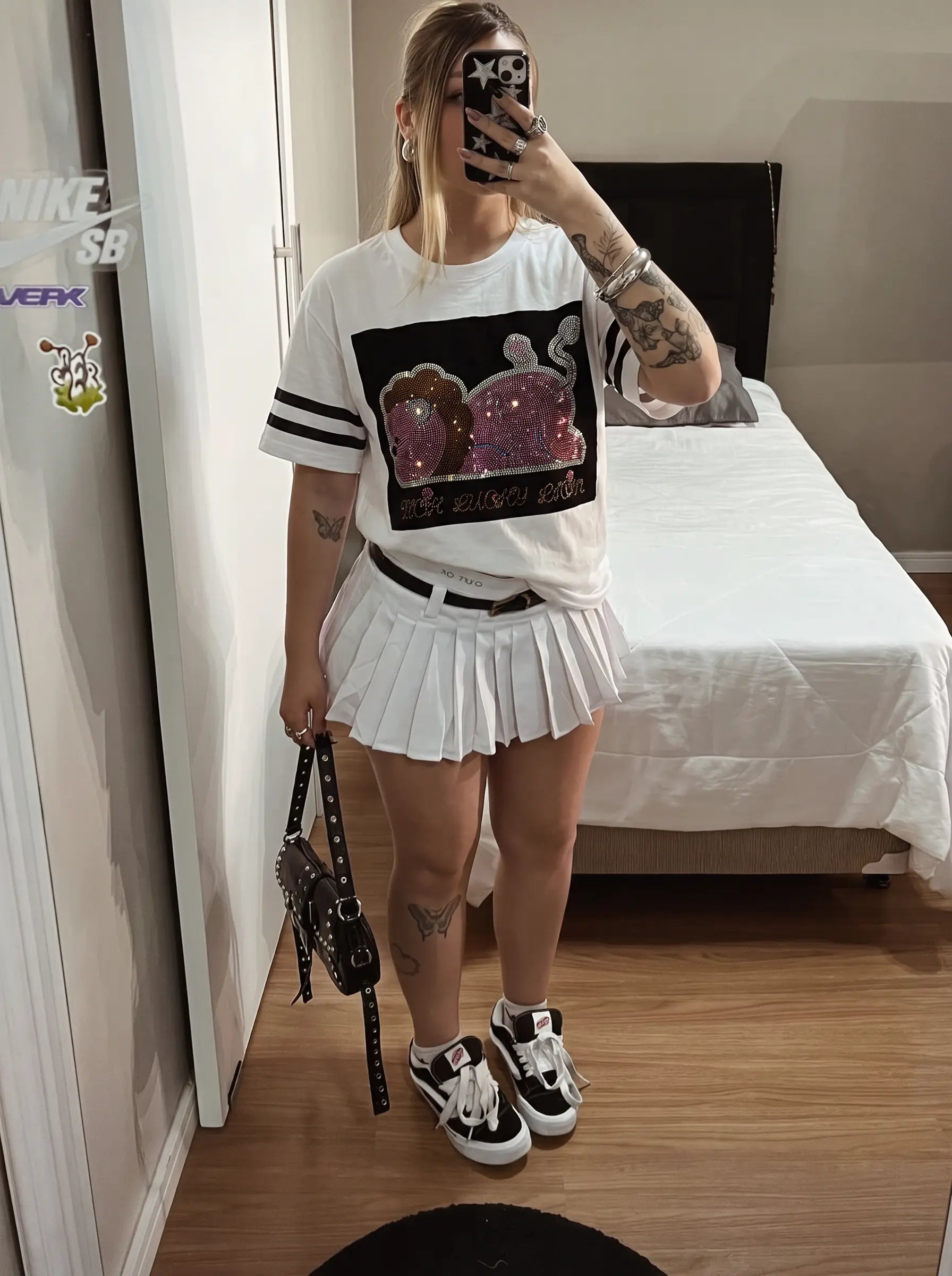 A woman takes a mirror selfie in a bedroom wearing a white graphic t-shirt with a black and pink sequined lion print, a white pleated mini skirt, and black and white sneakers. TCH Rhinestone Cartoon Pig Graphic Baseball T-Shirt