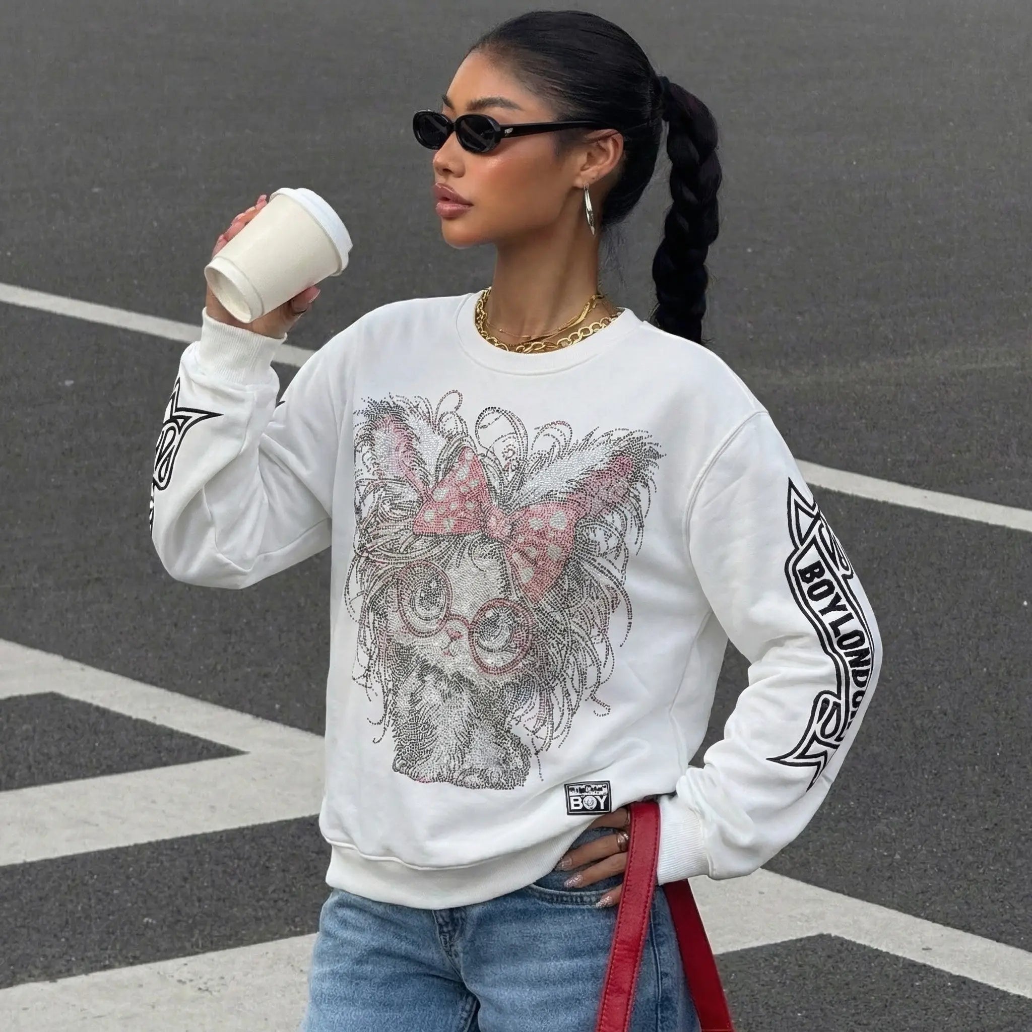 Woman wearing a white sweatshirt with a graphic design, holding a coffee cup on a street. TCH Rhinestone Cat Graphic Crew Neck Sweatshirt-2