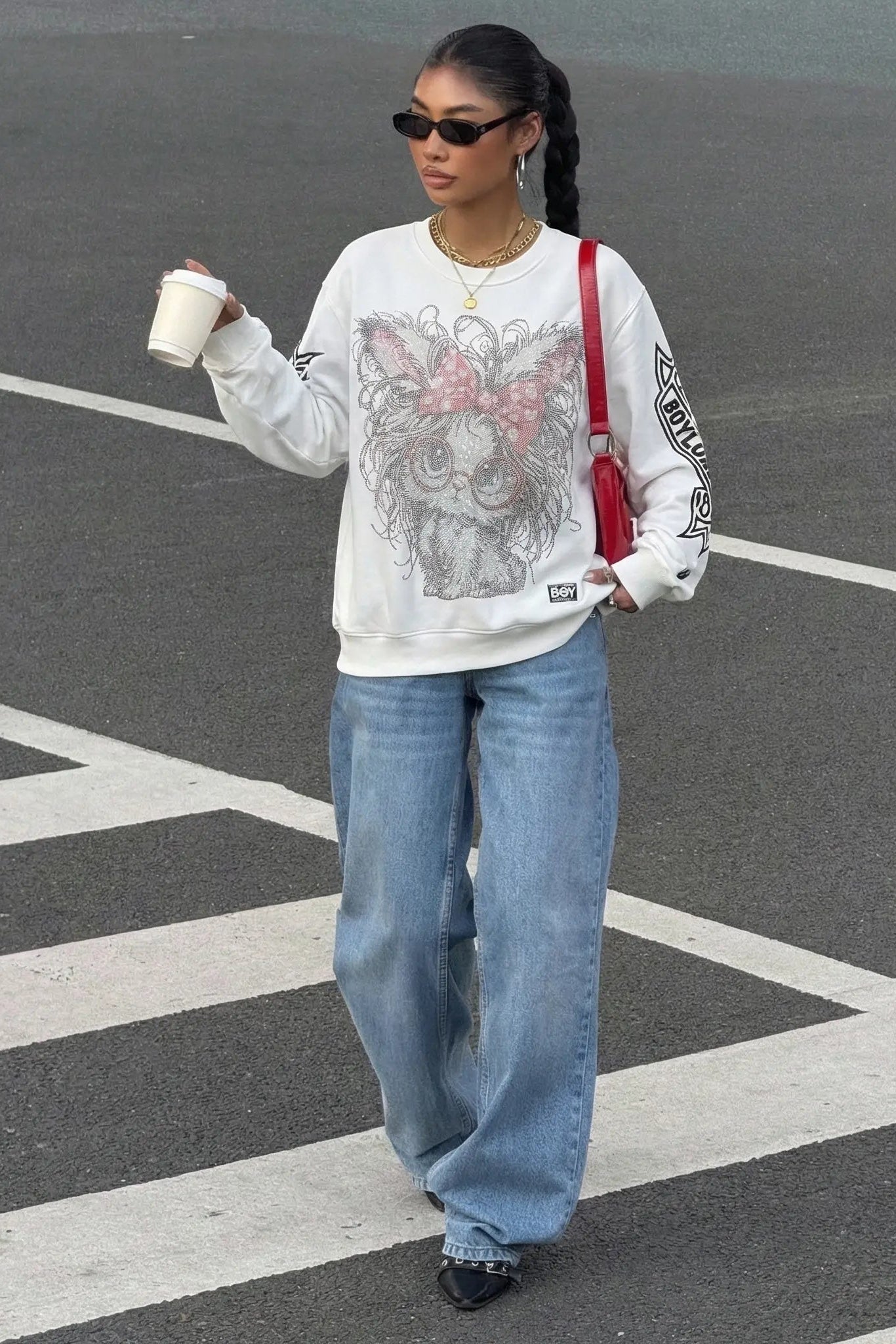 Woman walking on a street holding a coffee cup, wearing a graphic sweatshirt and jeans. TCH Rhinestone Cat Graphic Crew Neck Sweatshirt
