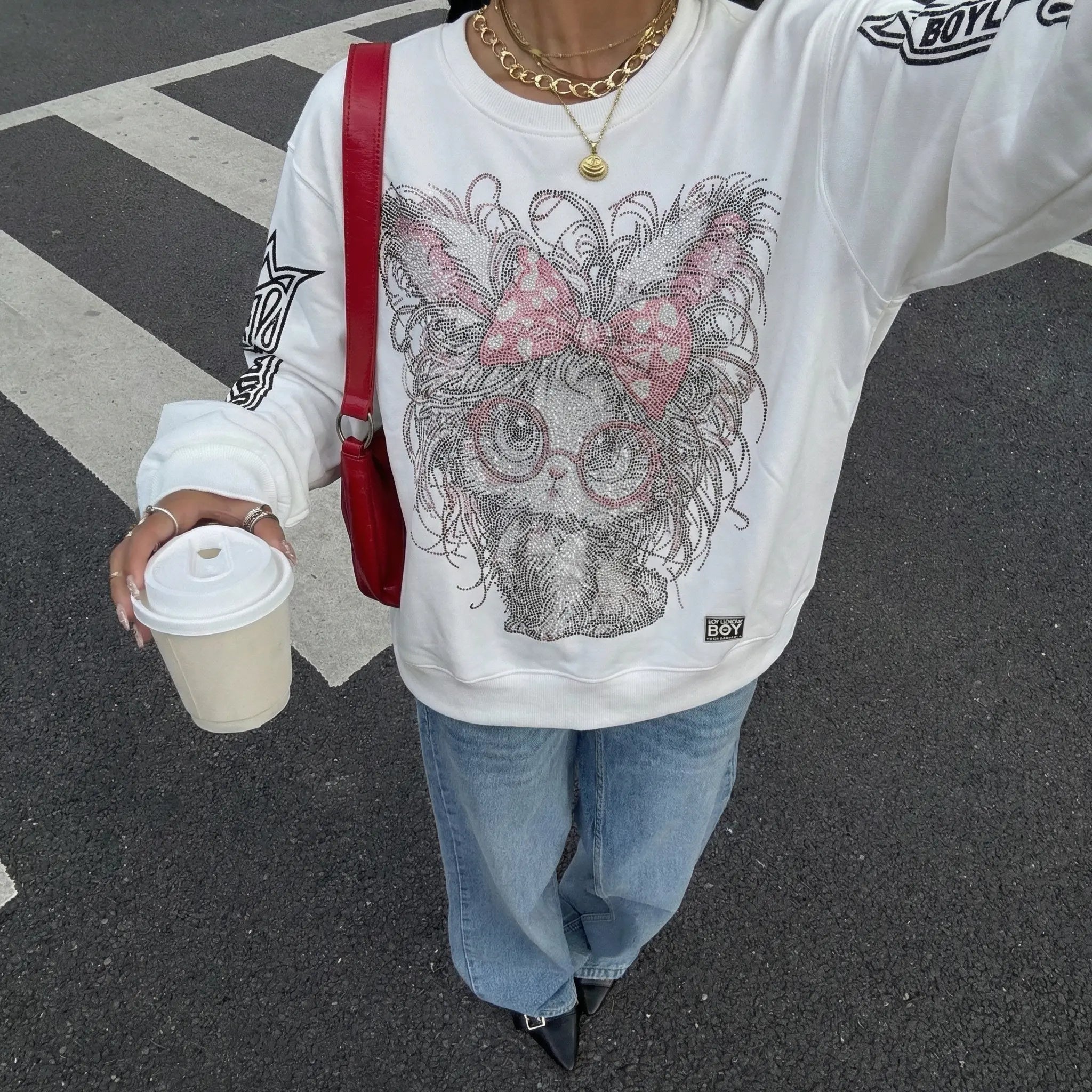 Person wearing a white sweatshirt with a graphic design, holding a coffee cup on a street. TCH Rhinestone Cat Graphic Crew Neck Sweatshirt