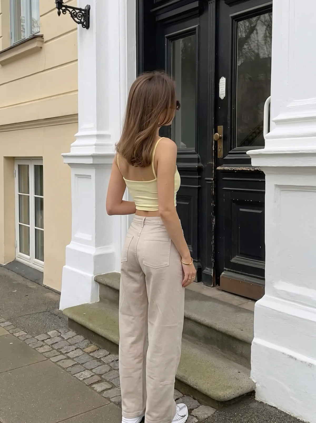 A woman stands with her back to the camera in front of a black door and white columns, wearing a pale yellow ribbed tank top and light beige wide-leg trousers. TCH Rhinestone Cherry Blossom Straight-Leg Jeans