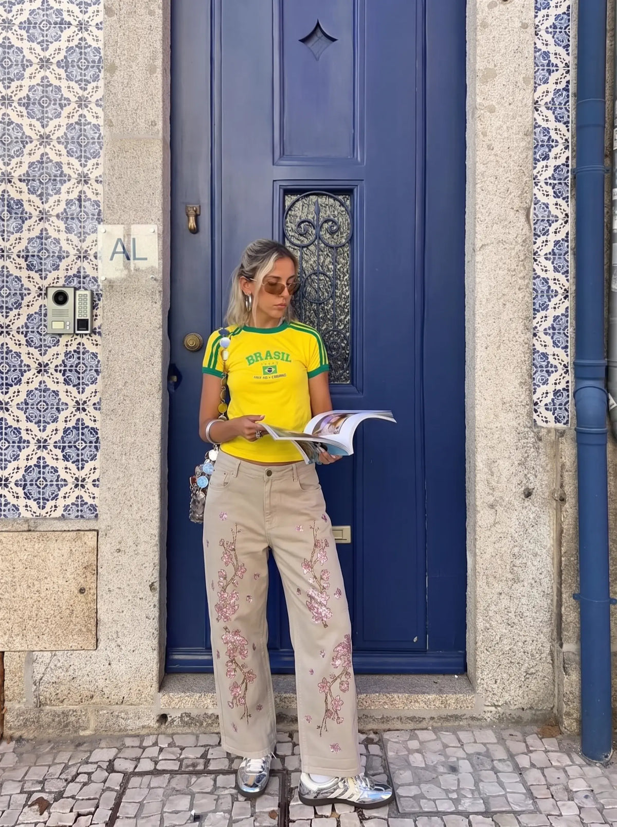 A woman stands in front of a blue door wearing a bright yellow t-shirt with green trim and 'BRASIL' printed on the chest, paired with beige wide-leg pants featuring pink floral embroidery. She is holding a magazine. TCH Rhinestone Cherry Blossom Straight-Leg Jeans