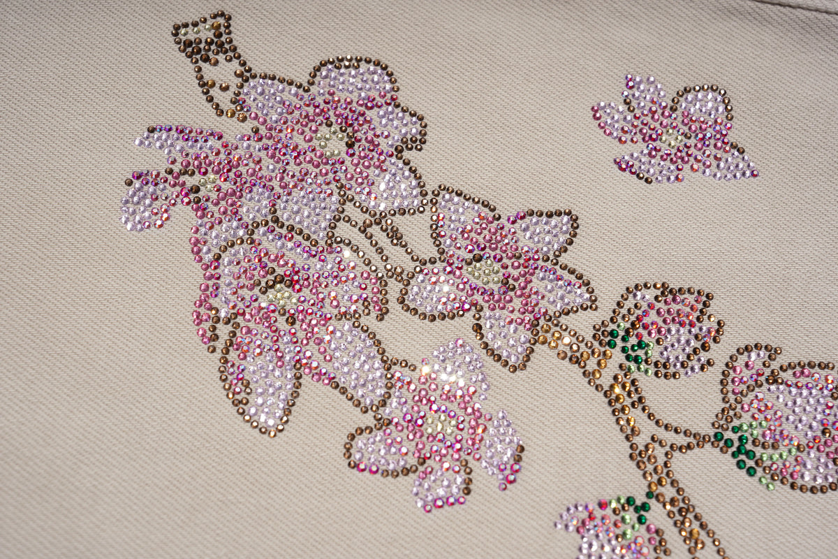 Close-up of a beige fabric surface decorated with pink and white beaded cherry blossom branches, featuring iridescent rhinestones and metallic gold outlines. TCH Rhinestone Cherry Blossom Straight-Leg Jeans