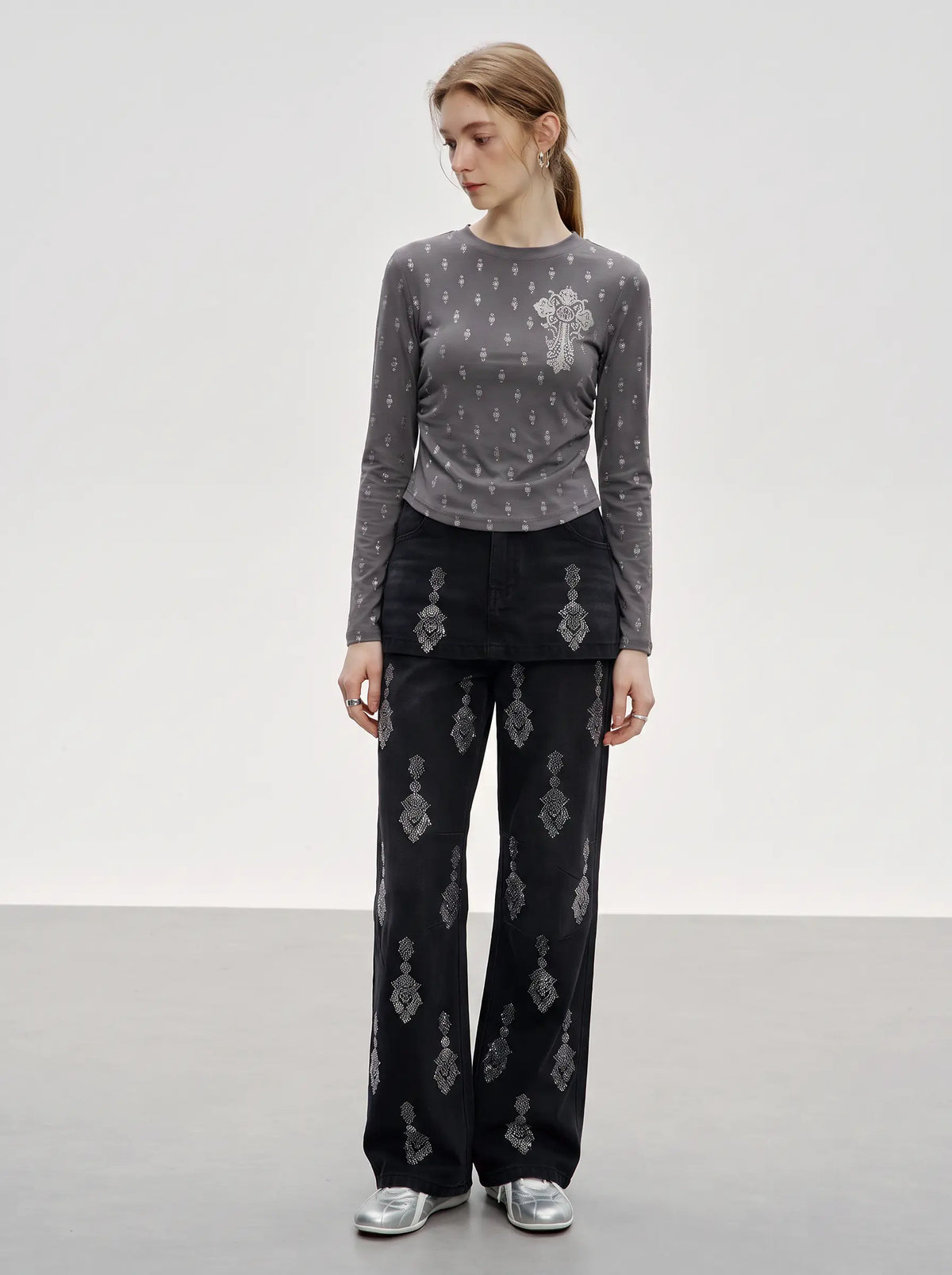 A model stands in a studio wearing a gray long-sleeve top with a small repeating pattern and a large floral applique on the chest, paired with black wide-leg pants featuring a larger repeating ornamental pattern, and silver shoes. TCH Rhinestone Cross Graphic Fitted Long Sleeve Top