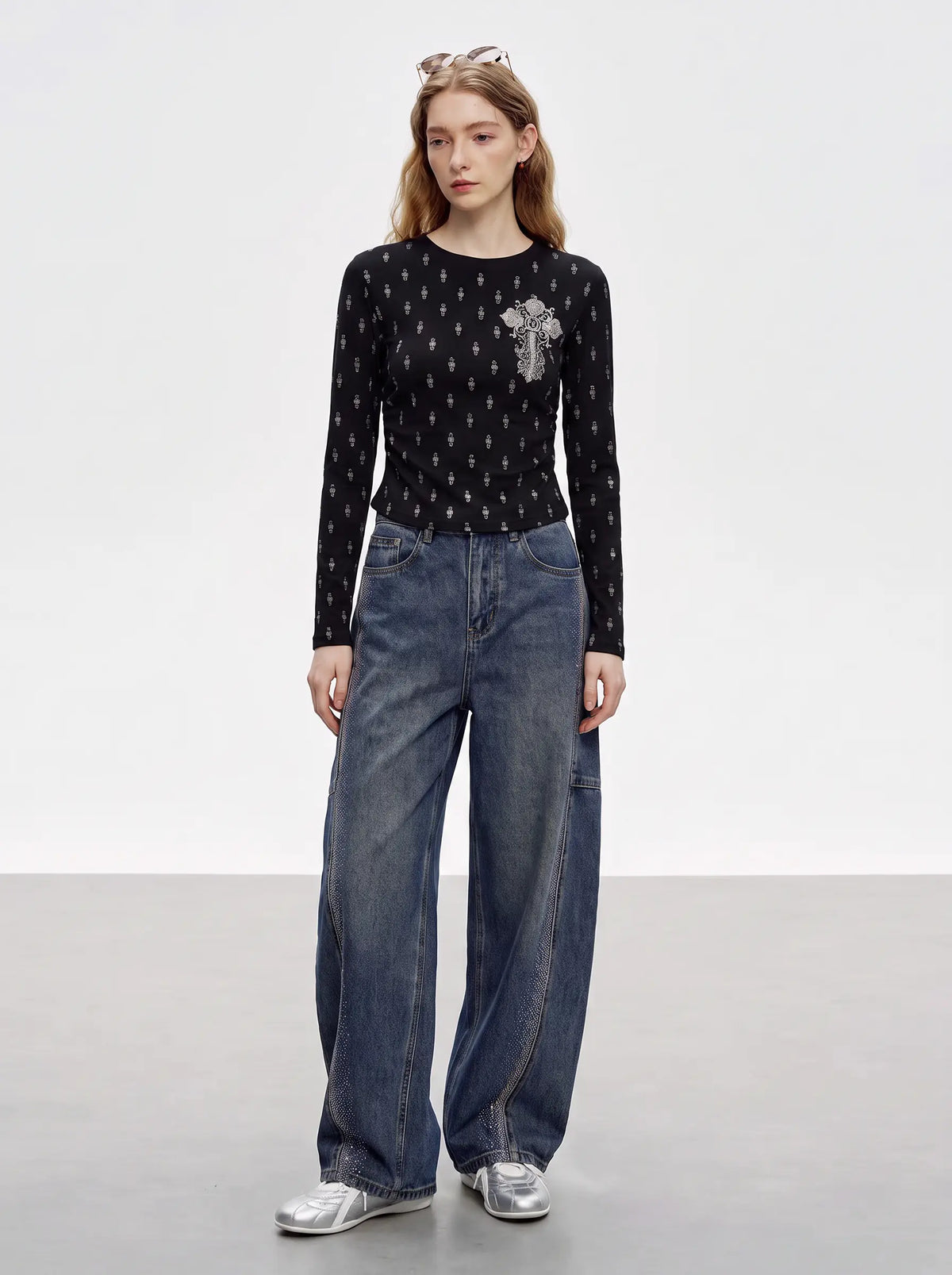A model stands in a studio wearing a black long-sleeve knit top with a small silver cross motif on the chest, paired with wide-leg blue jeans and silver sneakers. TCH Rhinestone Cross Graphic Fitted Long Sleeve Top