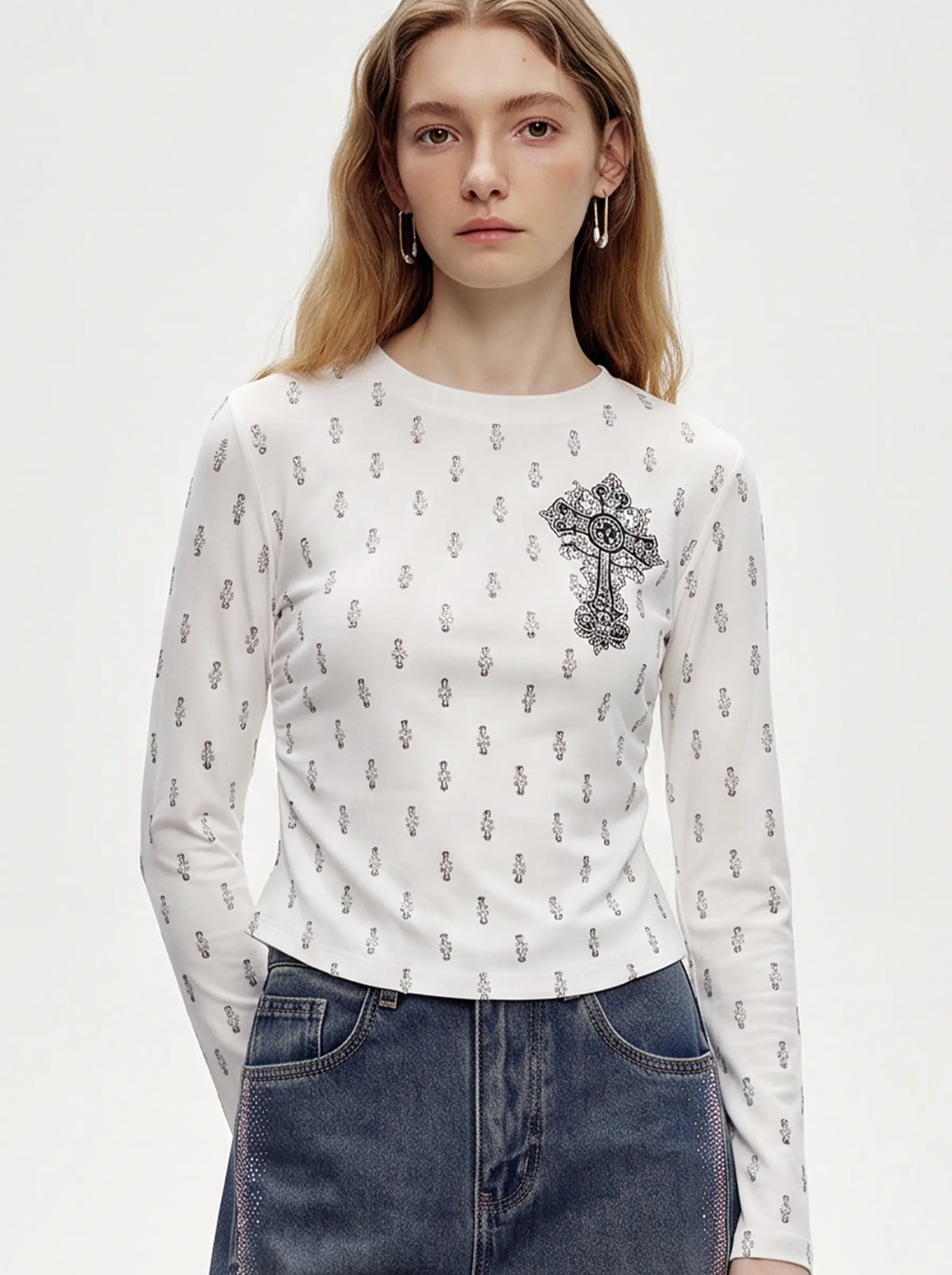 A woman wearing a white long-sleeve crewneck top with a small black repeating pattern and a large black ornate cross graphic on the left chest, paired with blue jeans against a light gray studio background. TCH Rhinestone Cross Graphic Fitted Long Sleeve Top