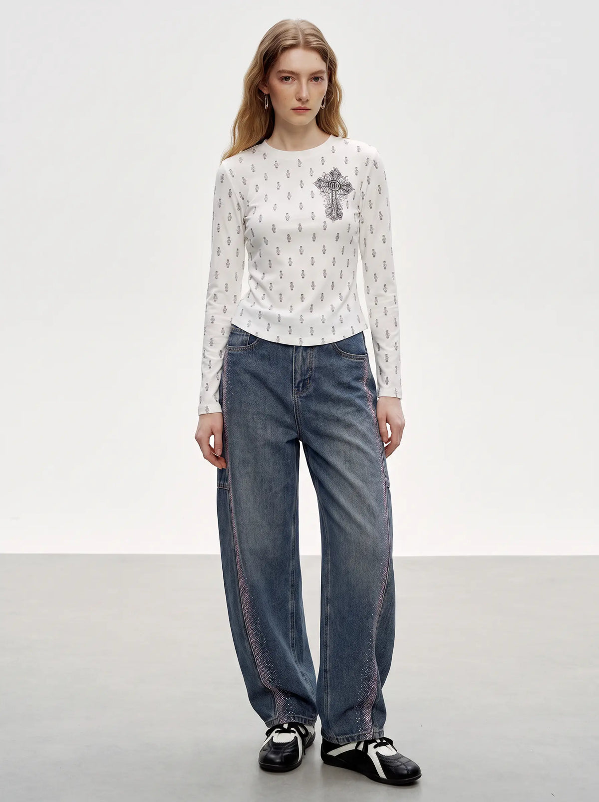 A model stands in a studio wearing a white long-sleeve top with a small gray cross graphic on the chest, paired with loose blue jeans and black-and-white sneakers. TCH Rhinestone Cross Graphic Fitted Long Sleeve Top