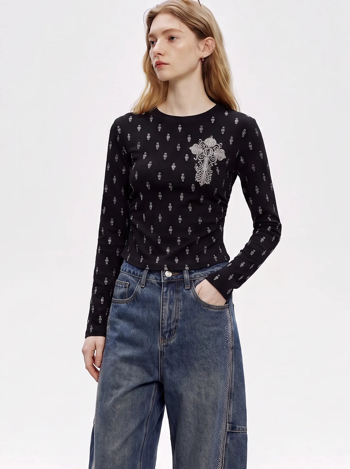 A woman modeling a black long-sleeve top with a small white cross pattern and a larger white cross embellishment on the chest, paired with high-waisted blue jeans against a plain white studio background. TCH Rhinestone Cross Graphic Fitted Long Sleeve Top