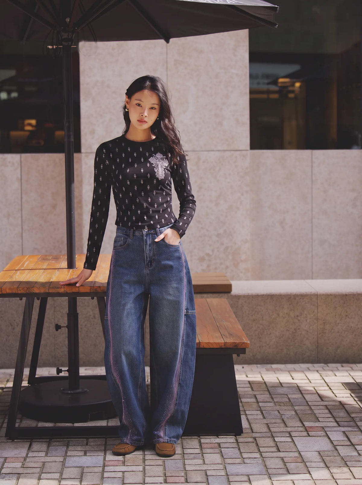 A woman stands outdoors next to a wooden table and bench under a black patio umbrella, wearing a black long-sleeve top with a white pattern and blue wide-leg jeans. TCH Rhinestone Cross Graphic Fitted Long Sleeve Top