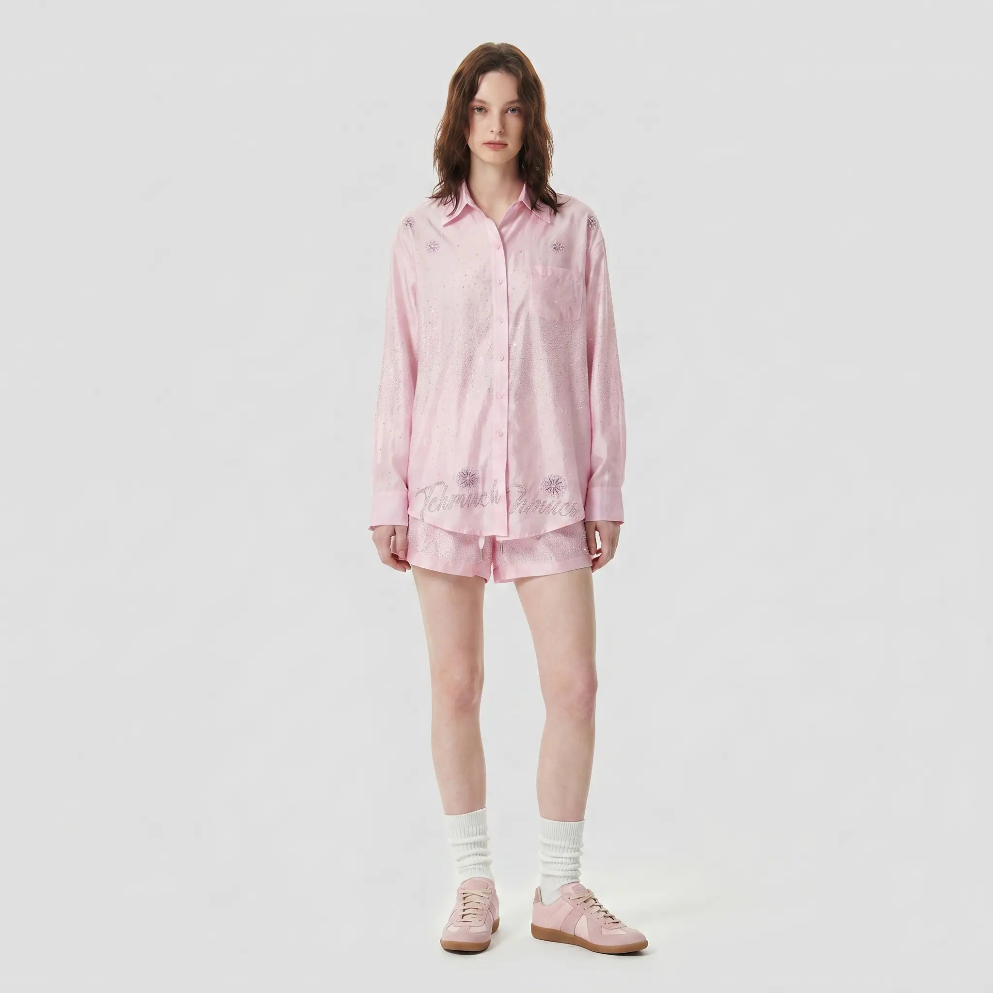 A model stands against a light gray studio background wearing a light pink long-sleeve button-up shirt dress with tonal floral embroidery, paired with white socks and pink shoes. TCH Rhinestone Crystal Embellished Pink Shirt & Shorts Set