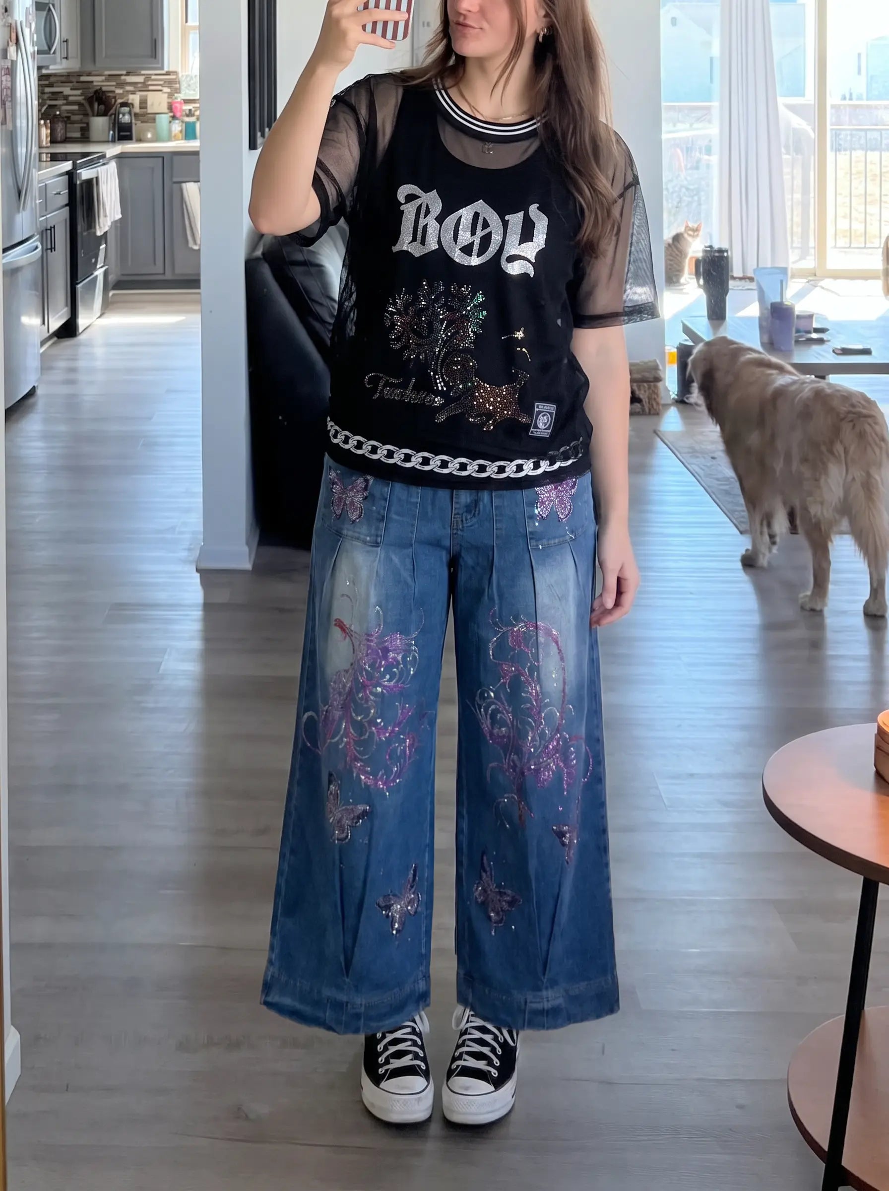 A woman takes a mirror selfie in a modern kitchen/dining room, wearing a black graphic t-shirt with 'BOY' in gothic lettering and a chain detail, paired with wide-leg blue jeans featuring purple and pink floral embroidery, and black-and-white sneakers. A golden retriever stands in the background. TCH Rhinestone Deer Graphic Mesh Sleeve Black Streetwear T-Shirt