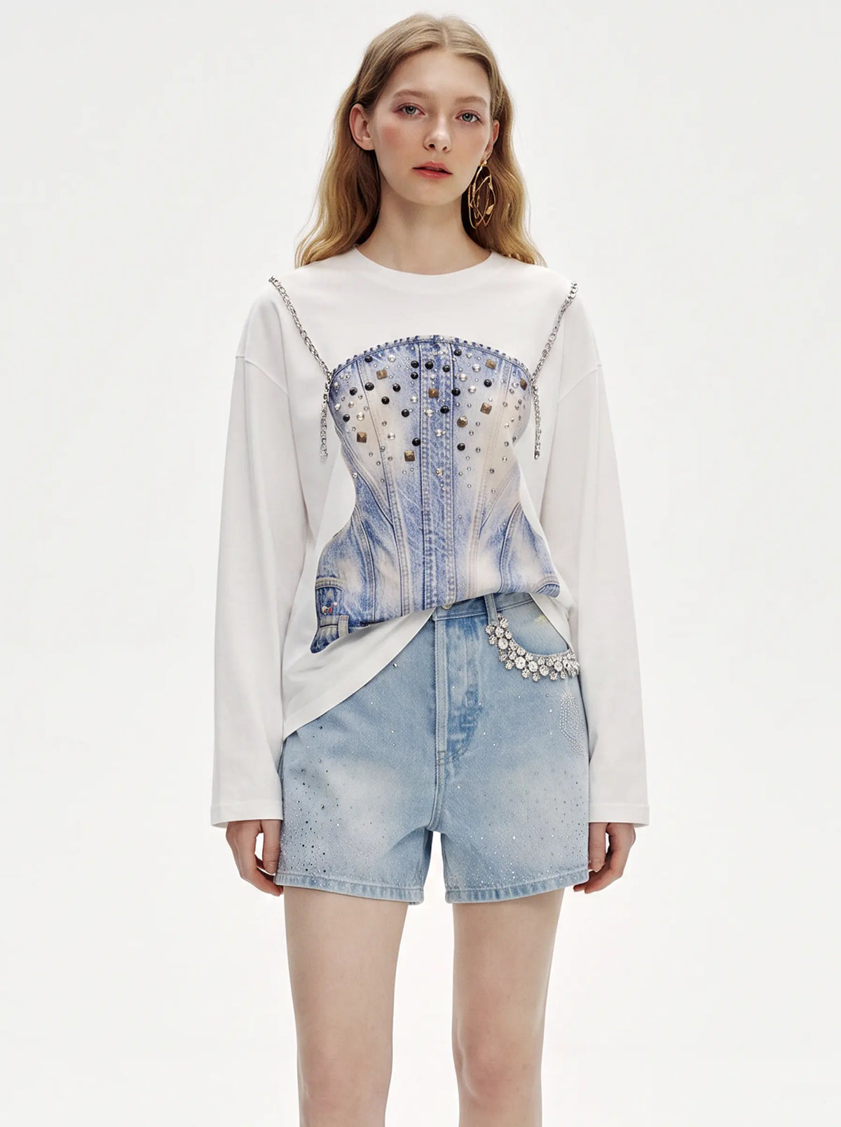 A model wearing a white long-sleeve top with a light blue denim corset overlay featuring silver studs and chain details, paired with light-wash denim shorts, against a plain white studio background. TCH Rhinestone Denim Corset Graphic Long Sleeve T-Shirt