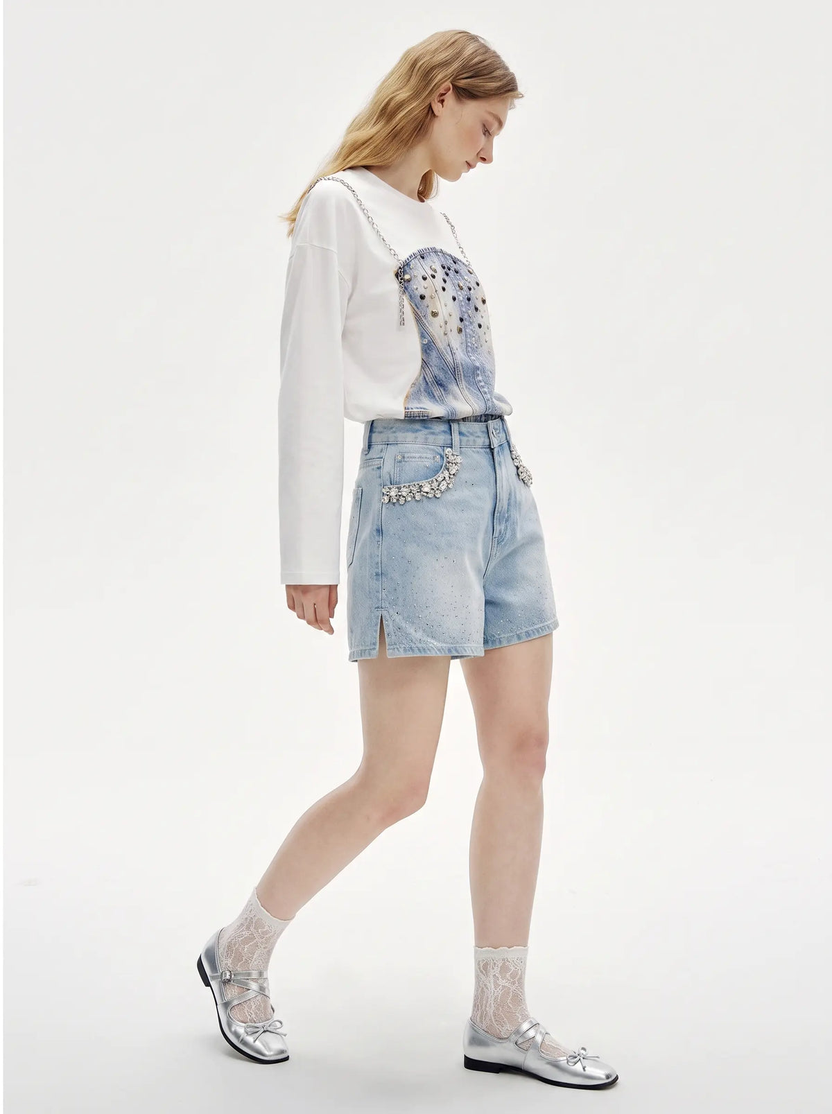 A model is shown from the side wearing a white long-sleeve top with a blue and silver graphic print, light-wash denim shorts with silver rhinestone embellishments, white lace socks, and silver loafers, against a plain white studio background. TCH Rhinestone Denim Corset Graphic Long Sleeve T-Shirt