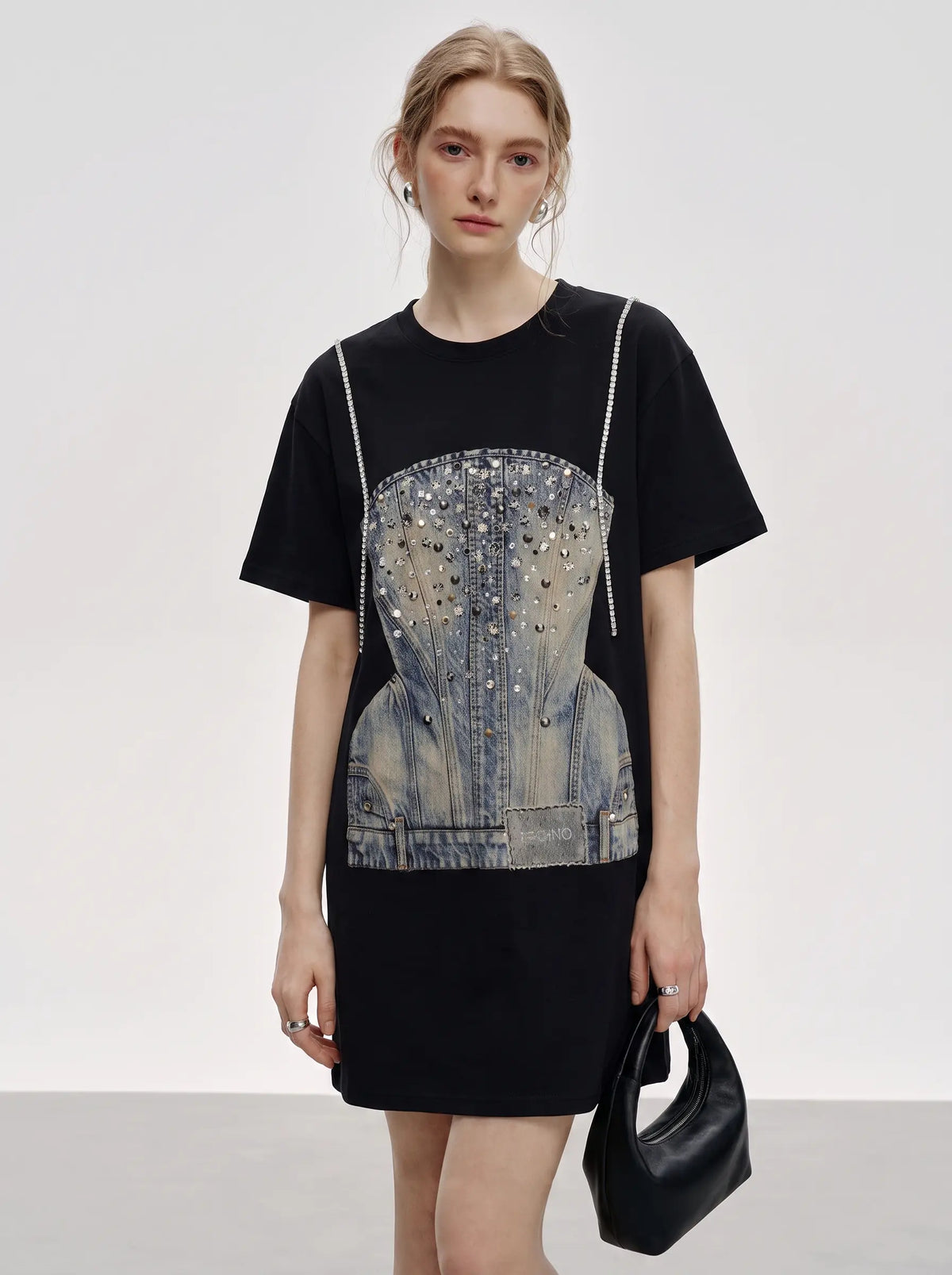 A model wearing a black short-sleeve t-shirt dress with a large denim corset graphic embellished with silver studs, holding a black leather handbag against a light gray studio background. TCH Rhinestone Denim Corset Graphic Oversized T-Shirt
