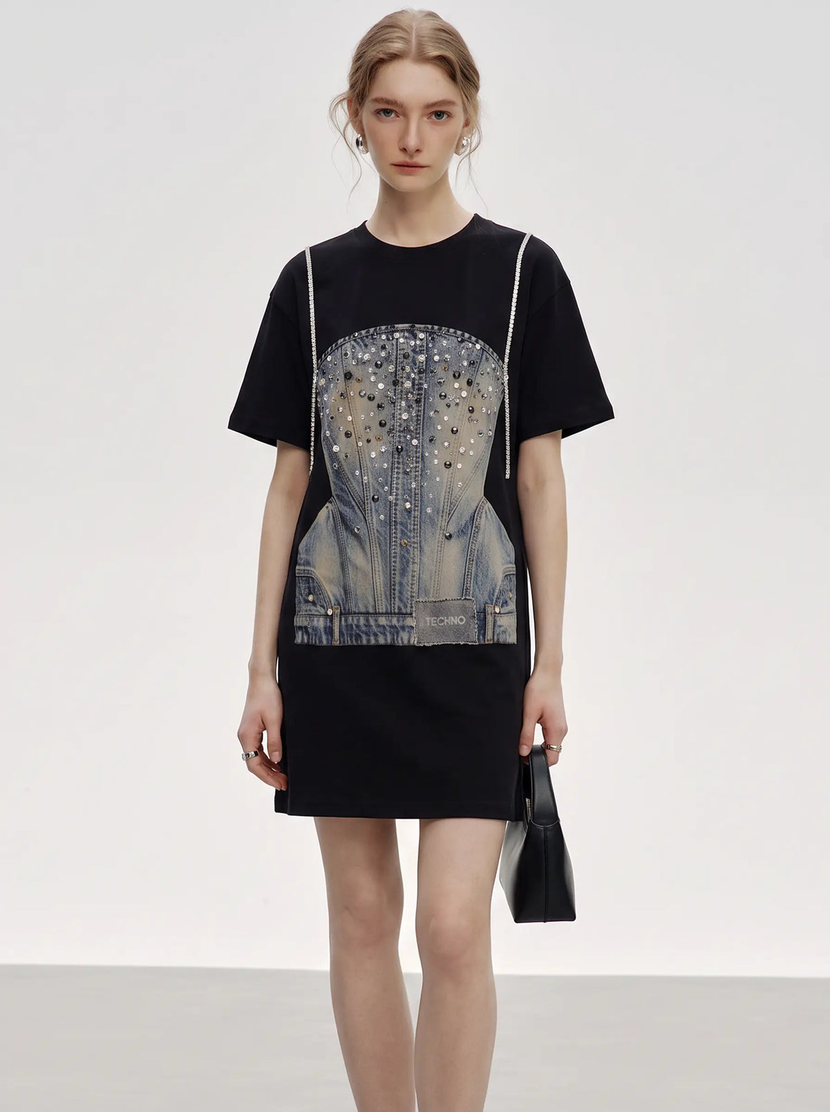 A model wearing a black short-sleeve mini dress with a large denim corset graphic embellished with rhinestones on the front, styled with a black handbag against a light gray studio background. TCH Rhinestone Denim Corset Graphic Oversized T-Shirt