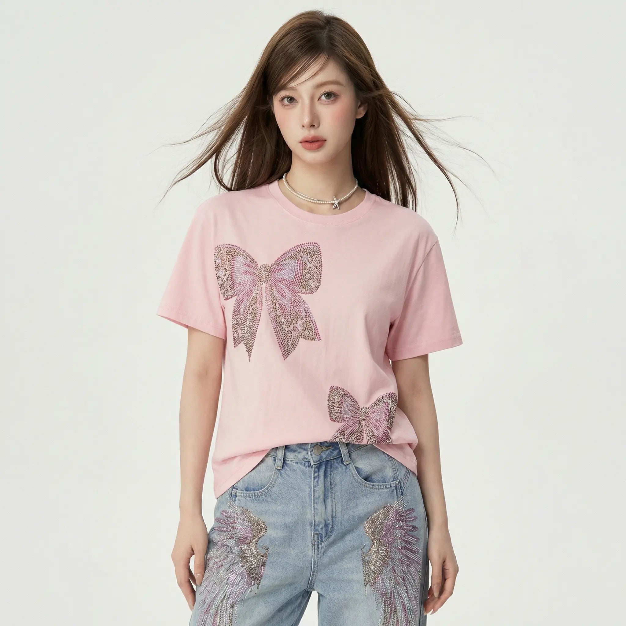 A woman modeling a light pink short-sleeve t-shirt with two large glittery bow designs on the front, paired with light blue jeans featuring angel wing embroidery, set against a plain off-white studio background. TCH Rhinestone Double Bow Cotton T-Shirt