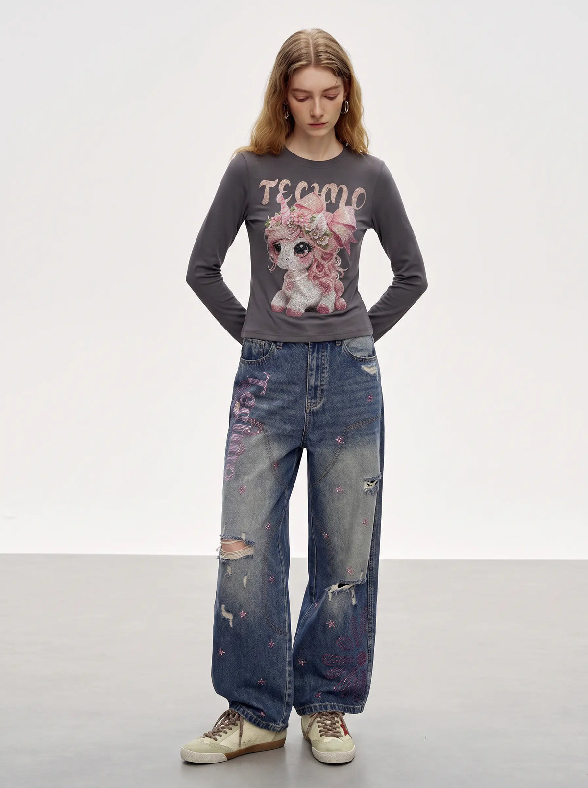 A model stands in a studio wearing a dark gray long-sleeve graphic t-shirt with a pink cartoon pony design and the word 'TECHNO' on the front, paired with distressed blue jeans and beige sneakers. TCH Rhinestone Fantasy Unicorn Graphic Long Sleeve T-Shirt