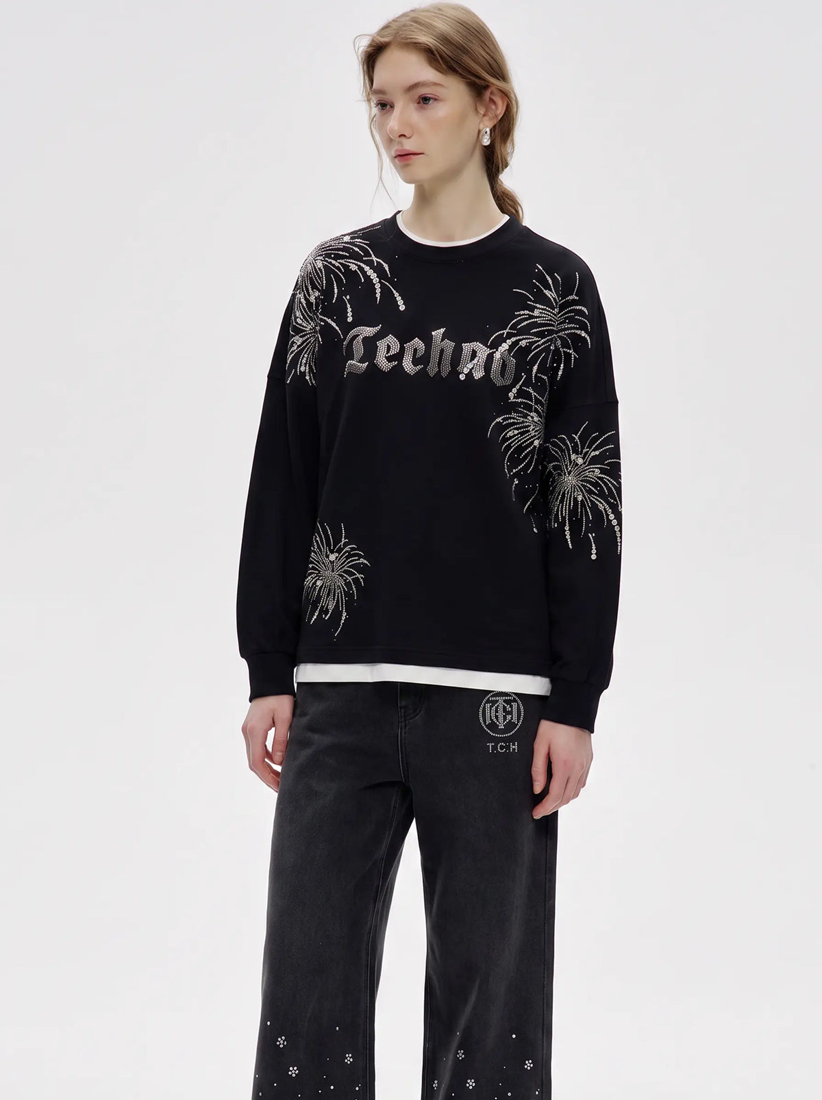 A model wearing a black crewneck sweatshirt with 'Techno' written in gothic letters and silver fireworks embellishments, paired with black jeans, against a plain light gray background. TCH Rhinestone Firework Graphic Black Sweatshirt