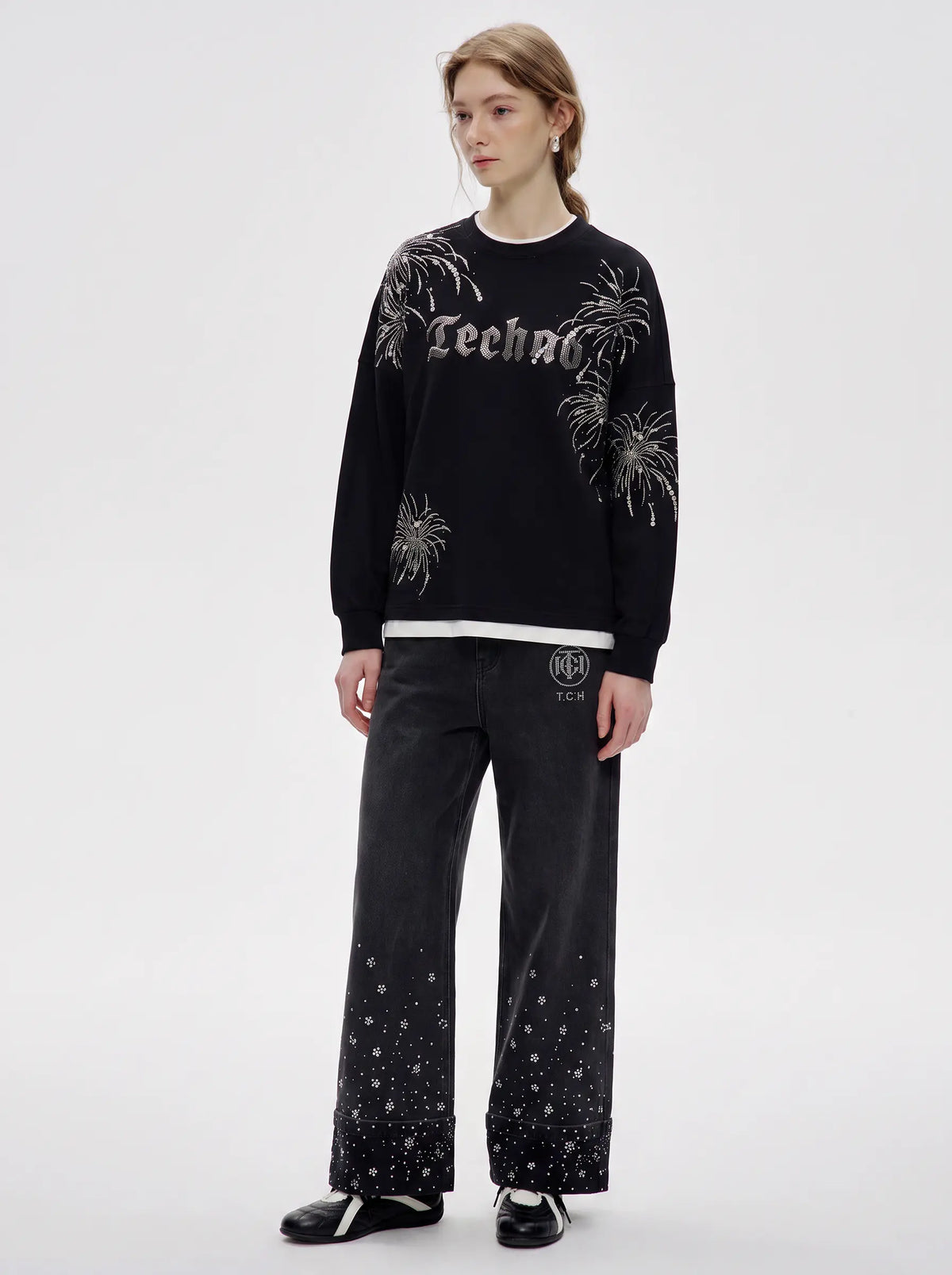 A model stands against a light gray studio background wearing a black crewneck sweatshirt with silver graphic embroidery, black wide-leg pants with silver speckled details, and black shoes. TCH Rhinestone Firework Graphic Black Sweatshirt