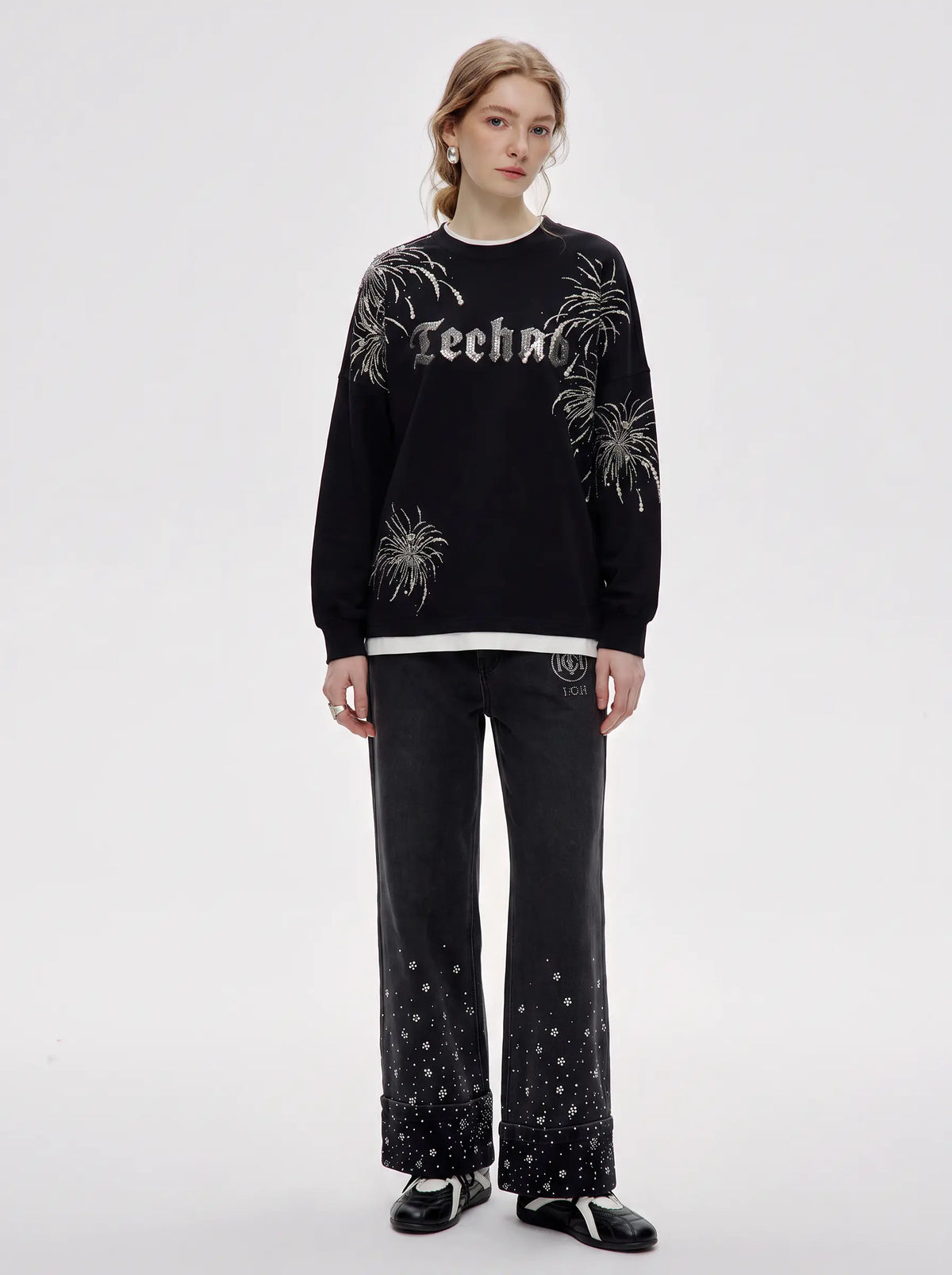 A model stands against a light gray studio background wearing a black crewneck sweatshirt with white graphic text and floral patterns, paired with black wide-leg pants featuring white speckled details near the hem, and black-and-white shoes. TCH Rhinestone Firework Graphic Black Sweatshirt