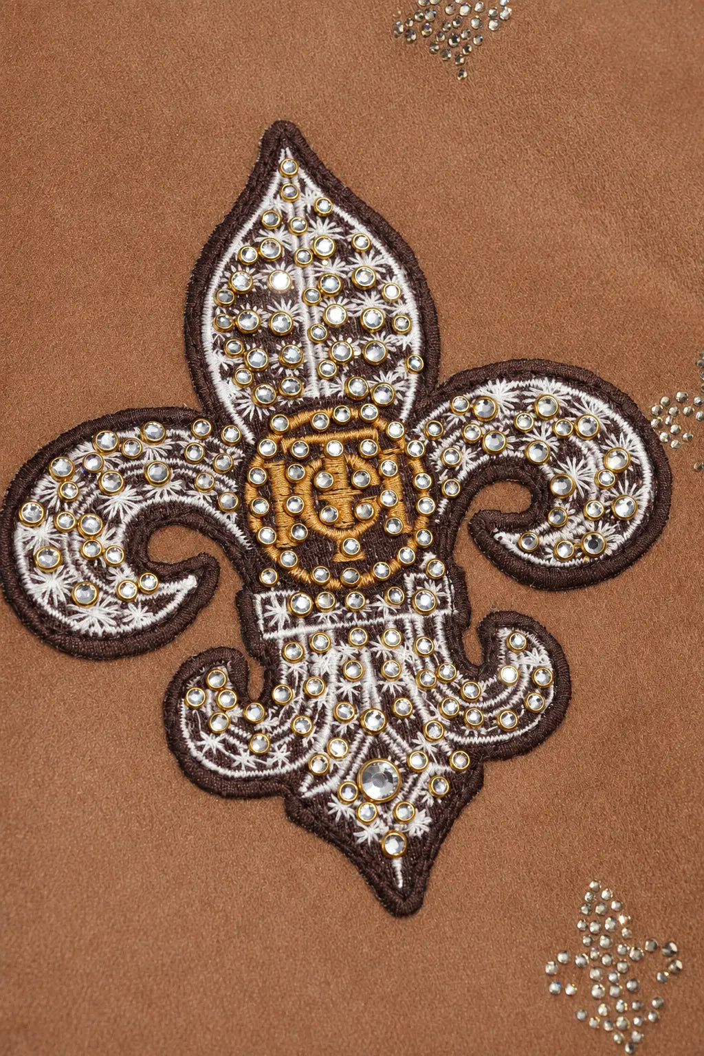 A close-up of a brown textured garment featuring a large embroidered fleur-de-lis patch with white floral embroidery, gold rhinestones, and a central gold monogram. TCH Rhinestone Fleur-de-Lis Hooded Jacket