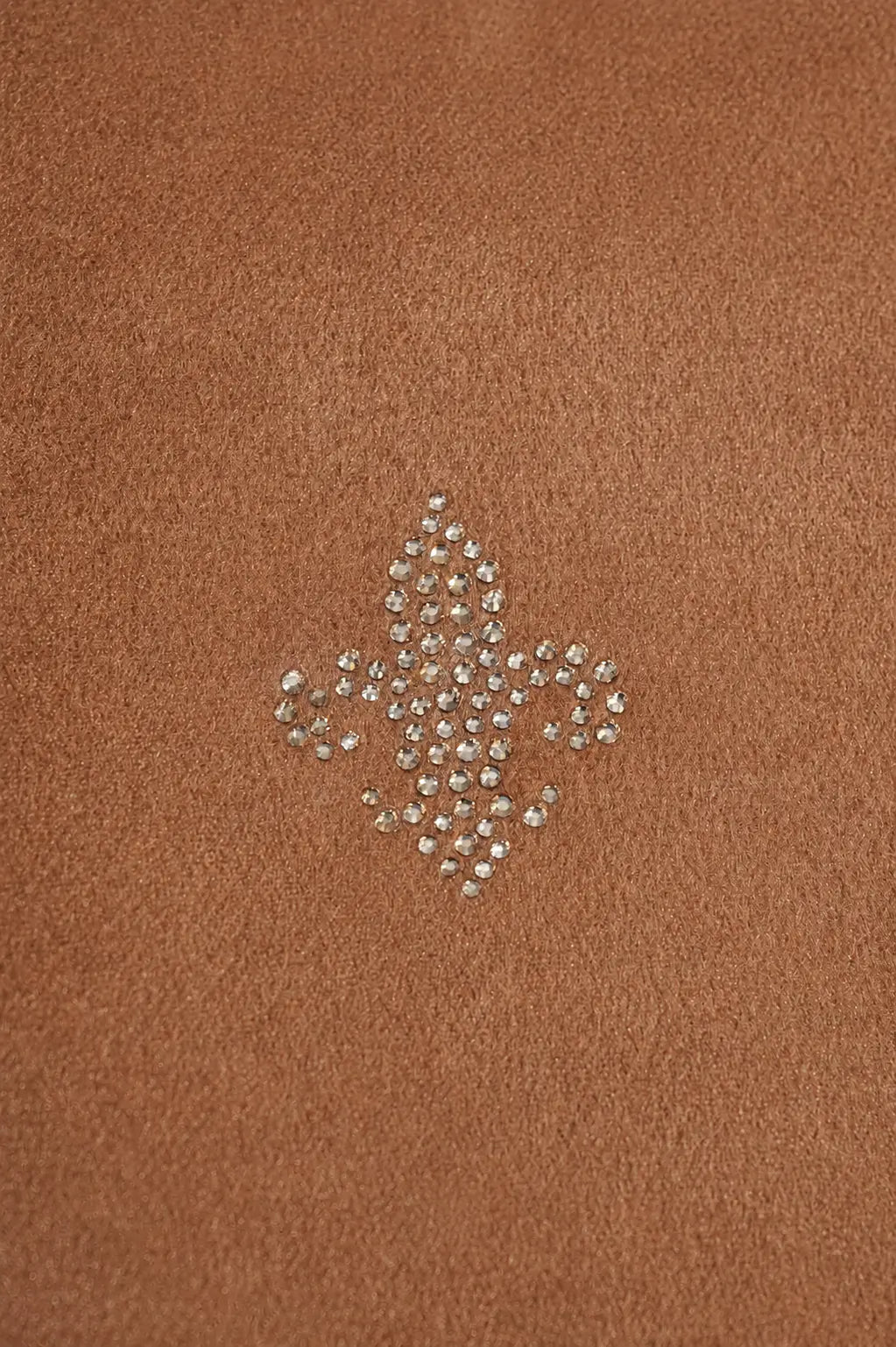 A close-up of a warm brown suede or faux-suede surface with a small rhinestone embellishment in the center forming a fleur-de-lis shape. TCH Rhinestone Fleur-de-Lis Hooded Jacket