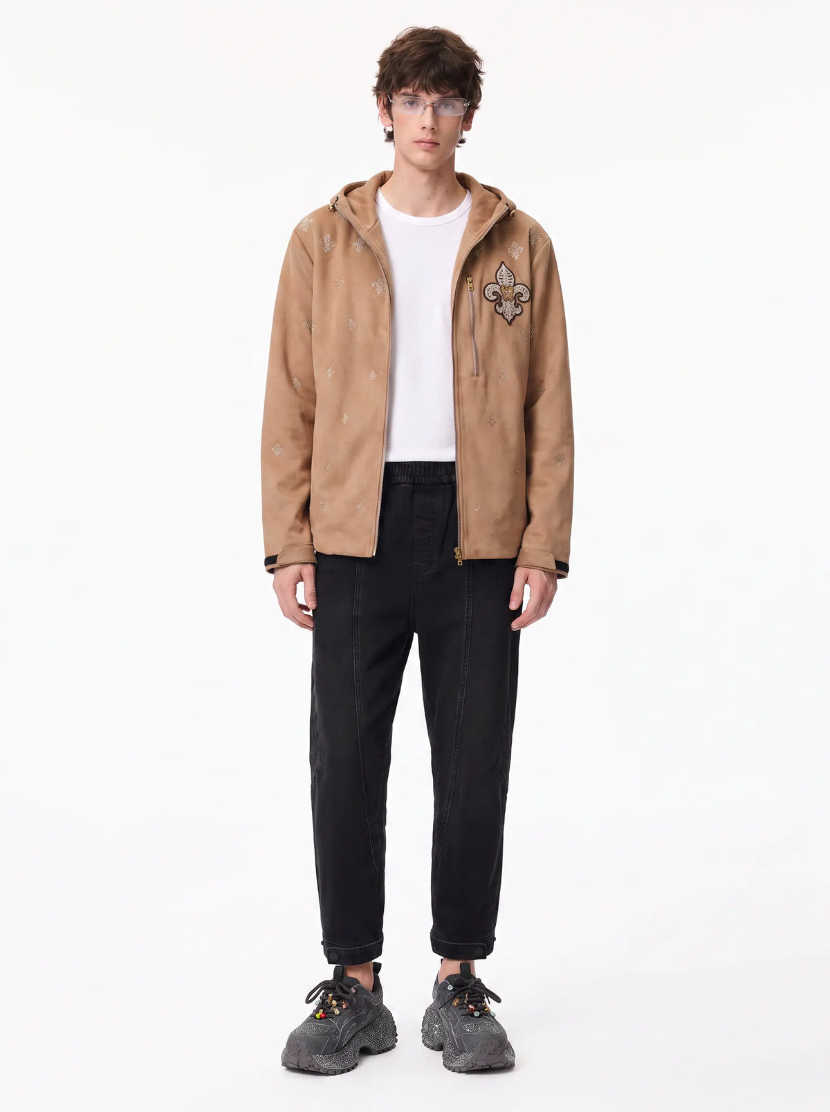 A model stands against a plain white studio background wearing a tan hooded jacket with a floral patch on the chest, a white t-shirt, black pants, and gray sneakers. TCH Rhinestone Fleur-de-Lis Hooded Jacket