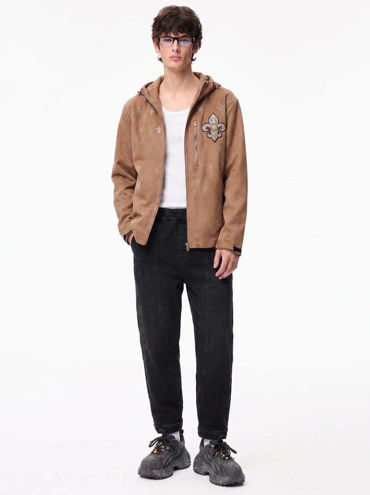 A model wearing a brown hooded jacket with a fleur-de-lis patch on the chest, styled with a white t-shirt, black jeans, and gray sneakers against a plain white background. TCH Rhinestone Fleur-de-Lis Hooded Jacket