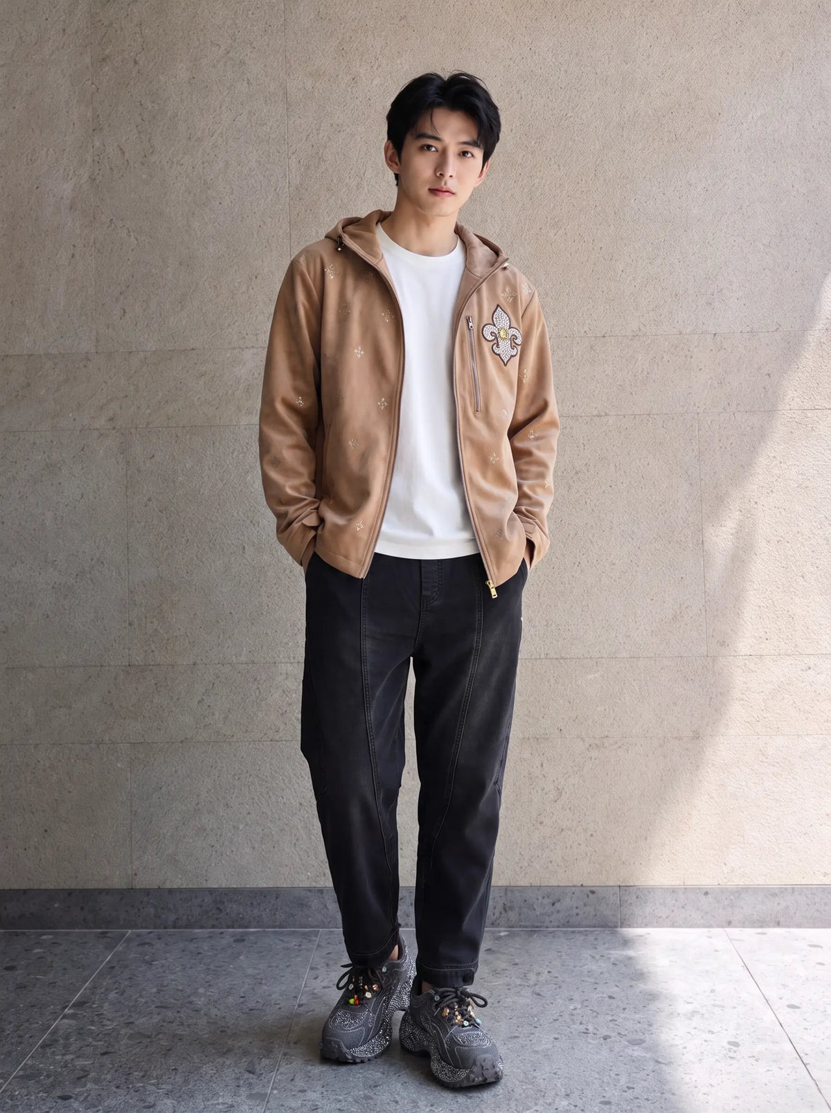 A man stands against a textured beige wall wearing a tan zip-up hoodie with a fox patch over a white t-shirt, dark charcoal pants, and gray sneakers. TCH Rhinestone Fleur-de-Lis Hooded Jacket