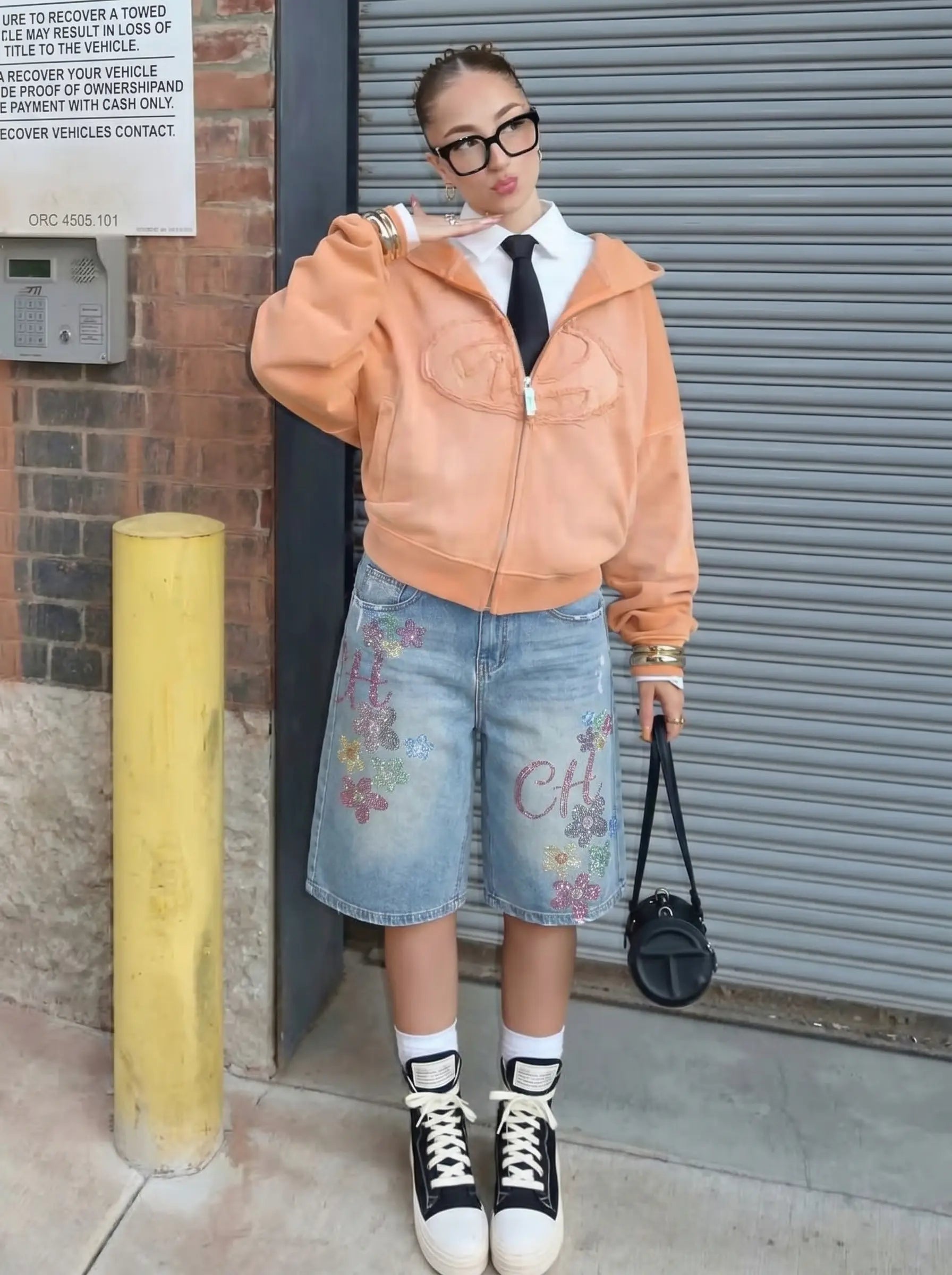 Person wearing a peach jacket, denim skirt, and black high-top sneakers standing against a metal shutter. TCH Rhinestone Floral Logo Wide-Leg Denim Shorts