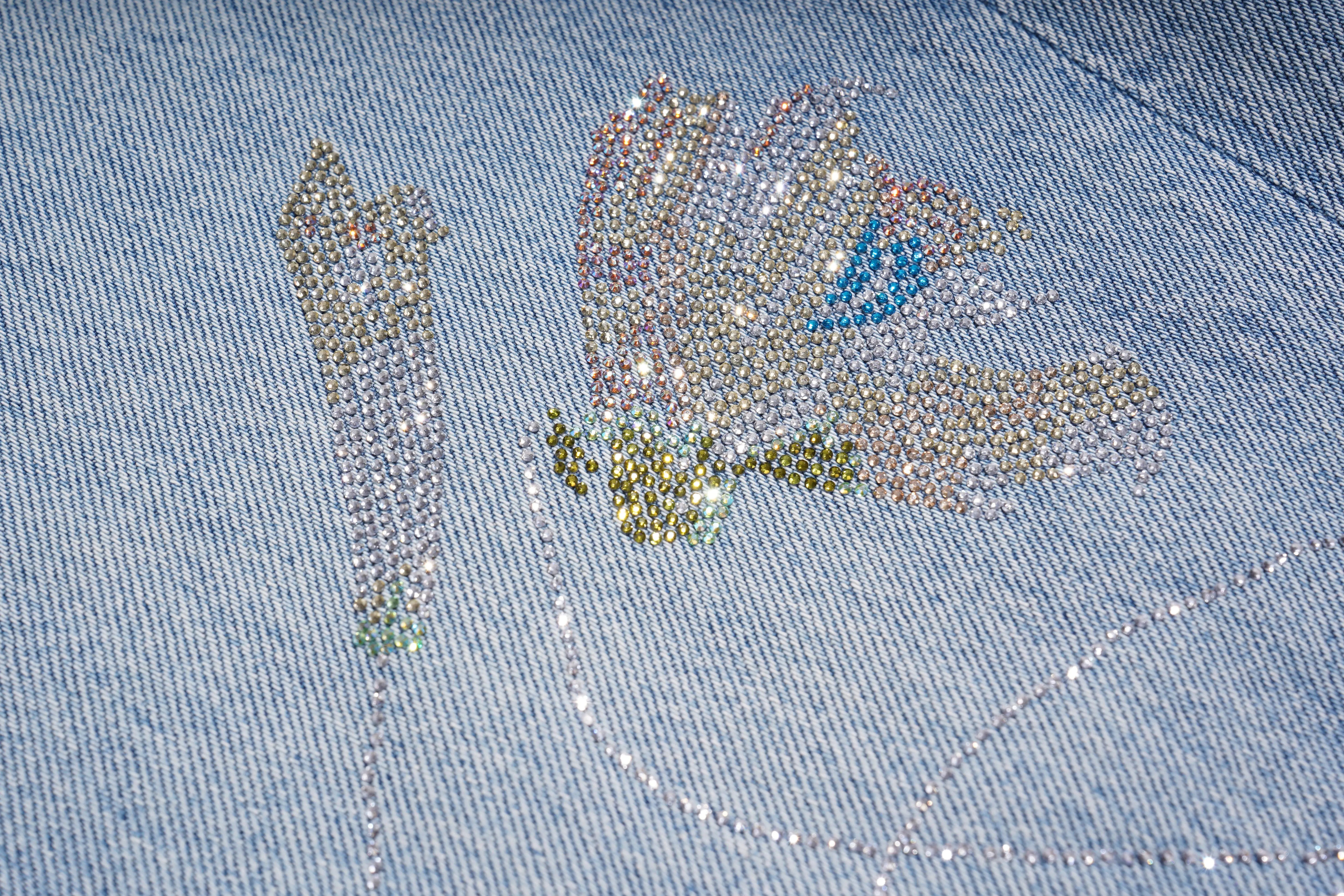 Close-up of a denim fabric with decorative lettering made from small stones or beads. TCH Rhinestone Floral Wide-Leg Blue Jeans