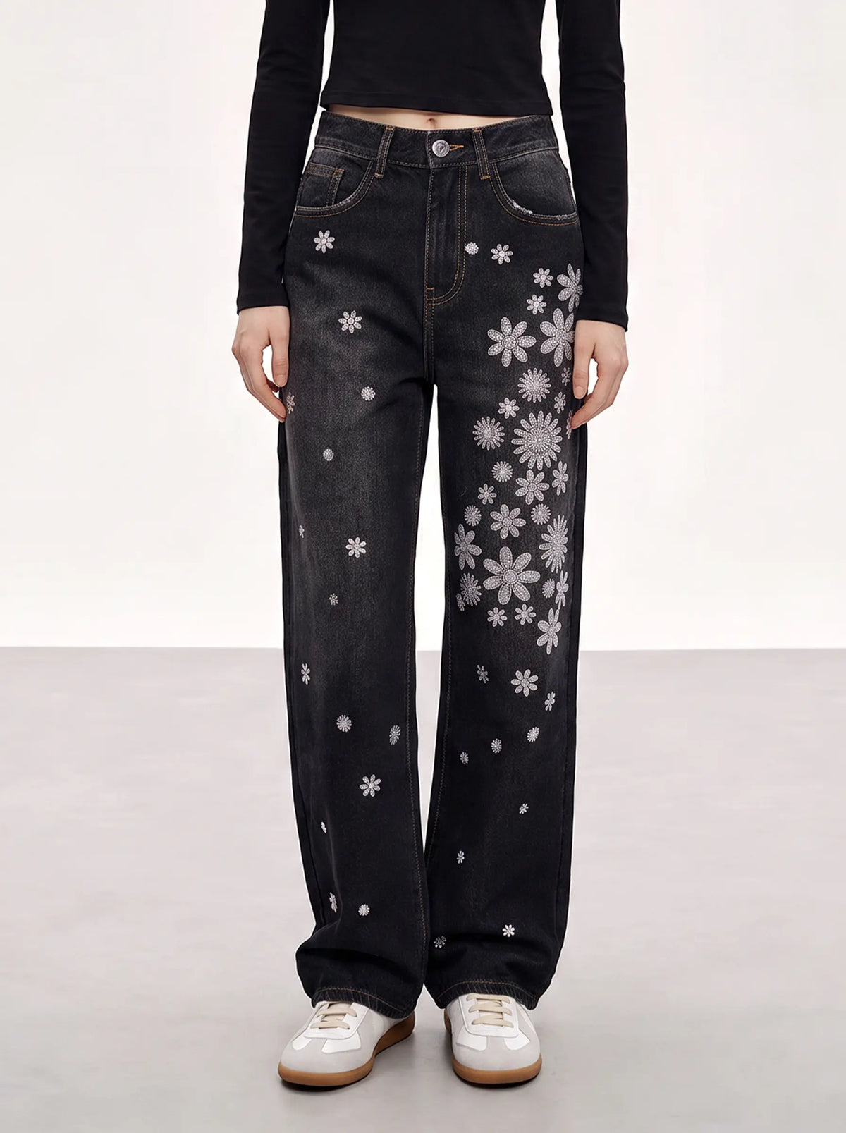 A model wearing black wide-leg jeans with white floral and snowflake patterns, paired with a black long-sleeve top and white sneakers, shown from the waist down against a neutral studio background. TCH Rhinestone Floral Wide-Leg Jeans
