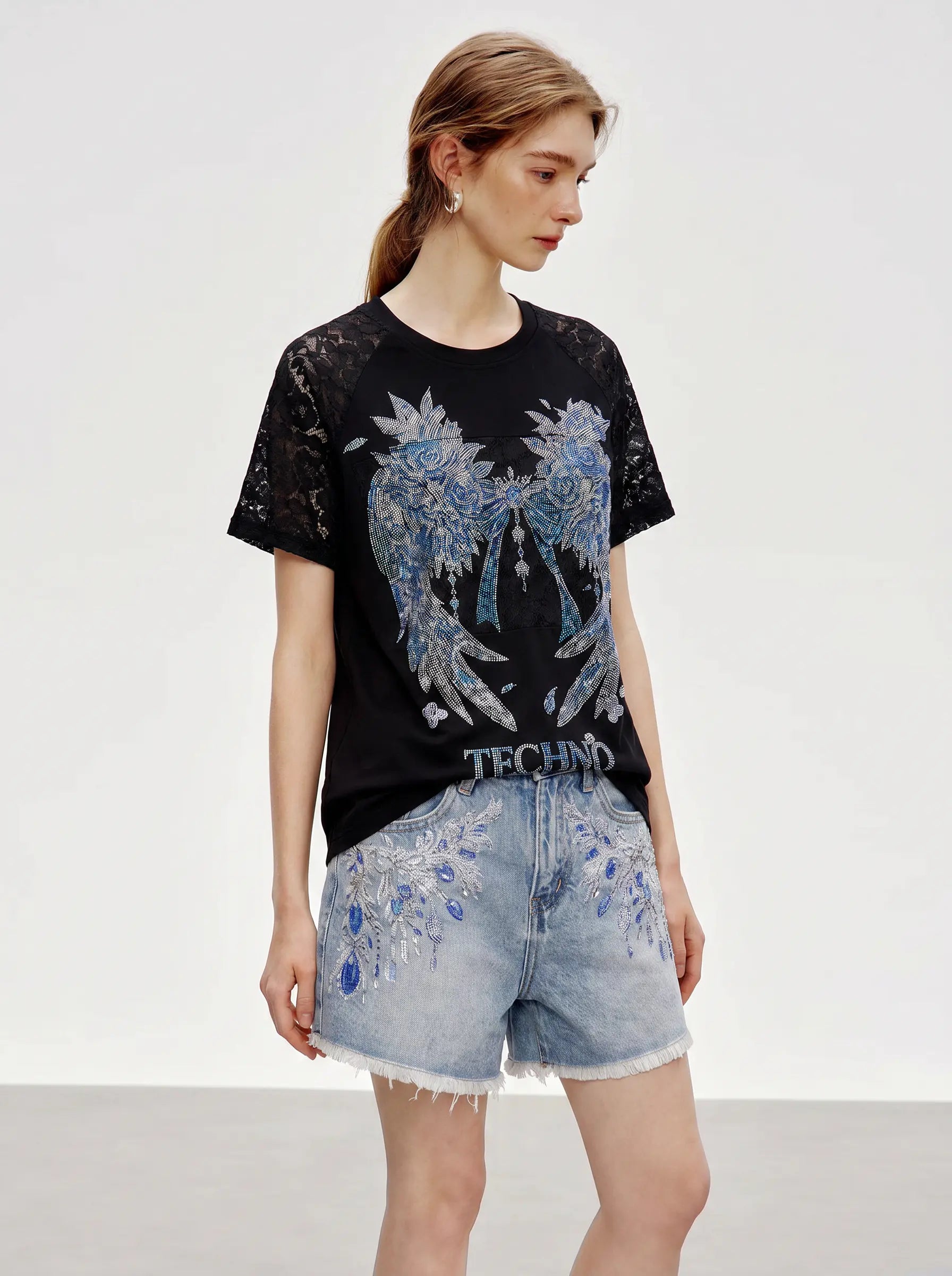 A model wearing a black short-sleeve t-shirt with a large blue and silver wing graphic and light blue denim shorts with matching blue embroidery, set against a plain light gray studio background. TCH Rhinestone Floral Wing Techno Graphic T-Shirt