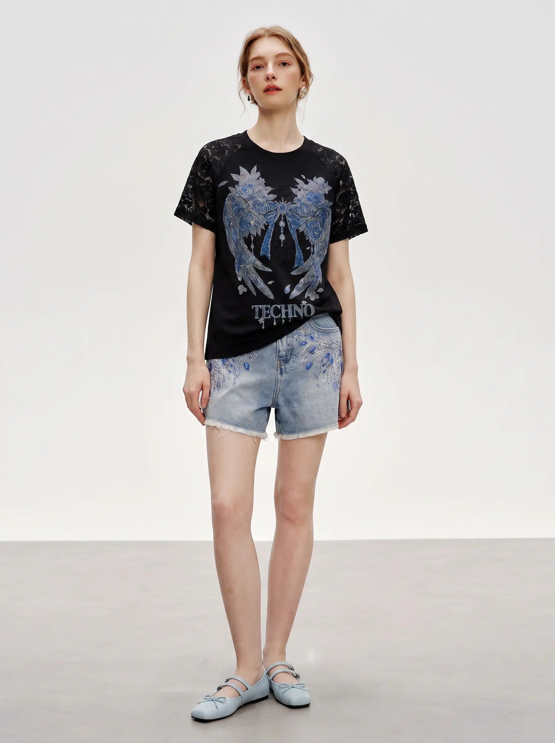 A model stands in a studio wearing a black short-sleeve graphic T-shirt with a large blue and white winged design and the word 'TECHNO' on the front, paired with light blue denim shorts and pale blue flats. TCH Rhinestone Floral Wing Techno Graphic T-Shirt