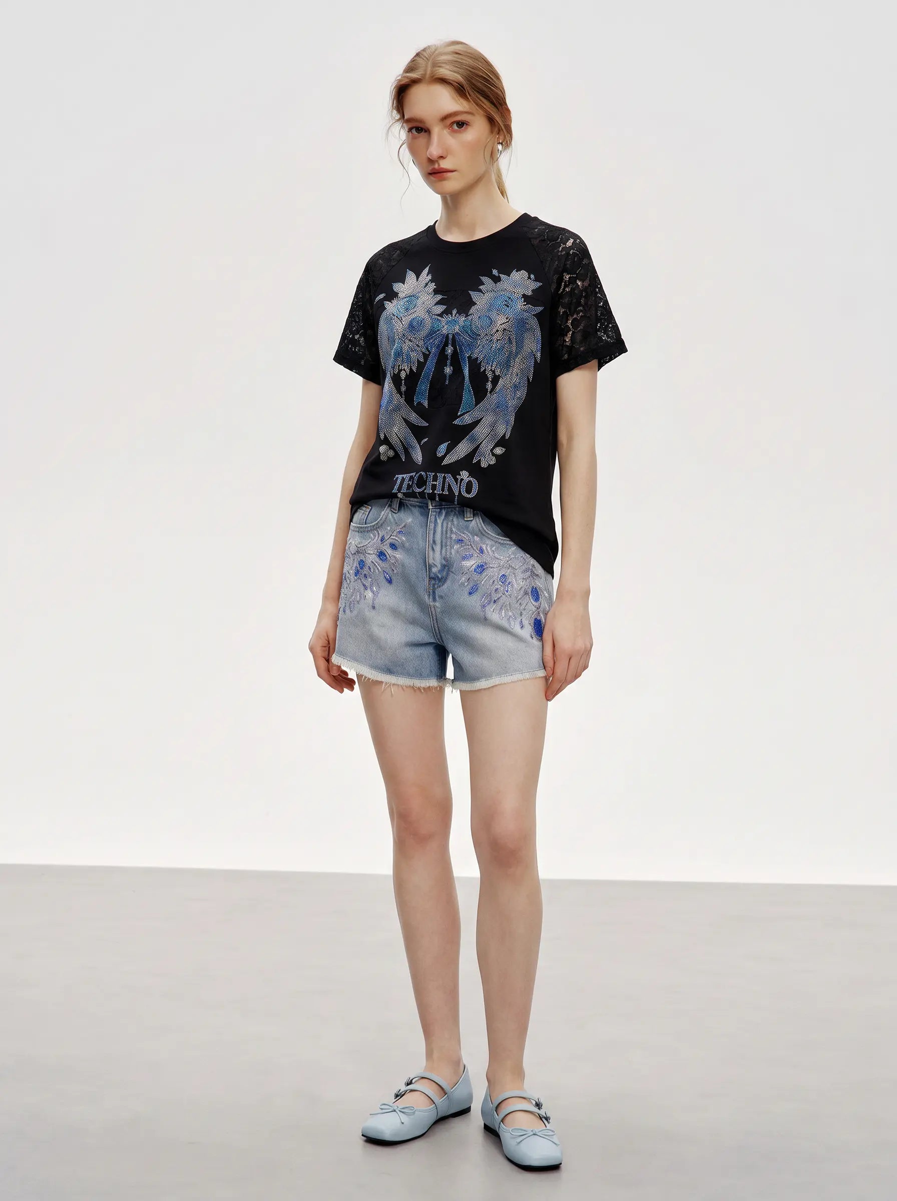 A model stands in a studio wearing a black short-sleeve graphic T-shirt with a large blue and white winged design and the word 'TECHNO' on the front, paired with light blue denim shorts and matching light blue flats. TCH Rhinestone Floral Wing Techno Graphic T-Shirt
