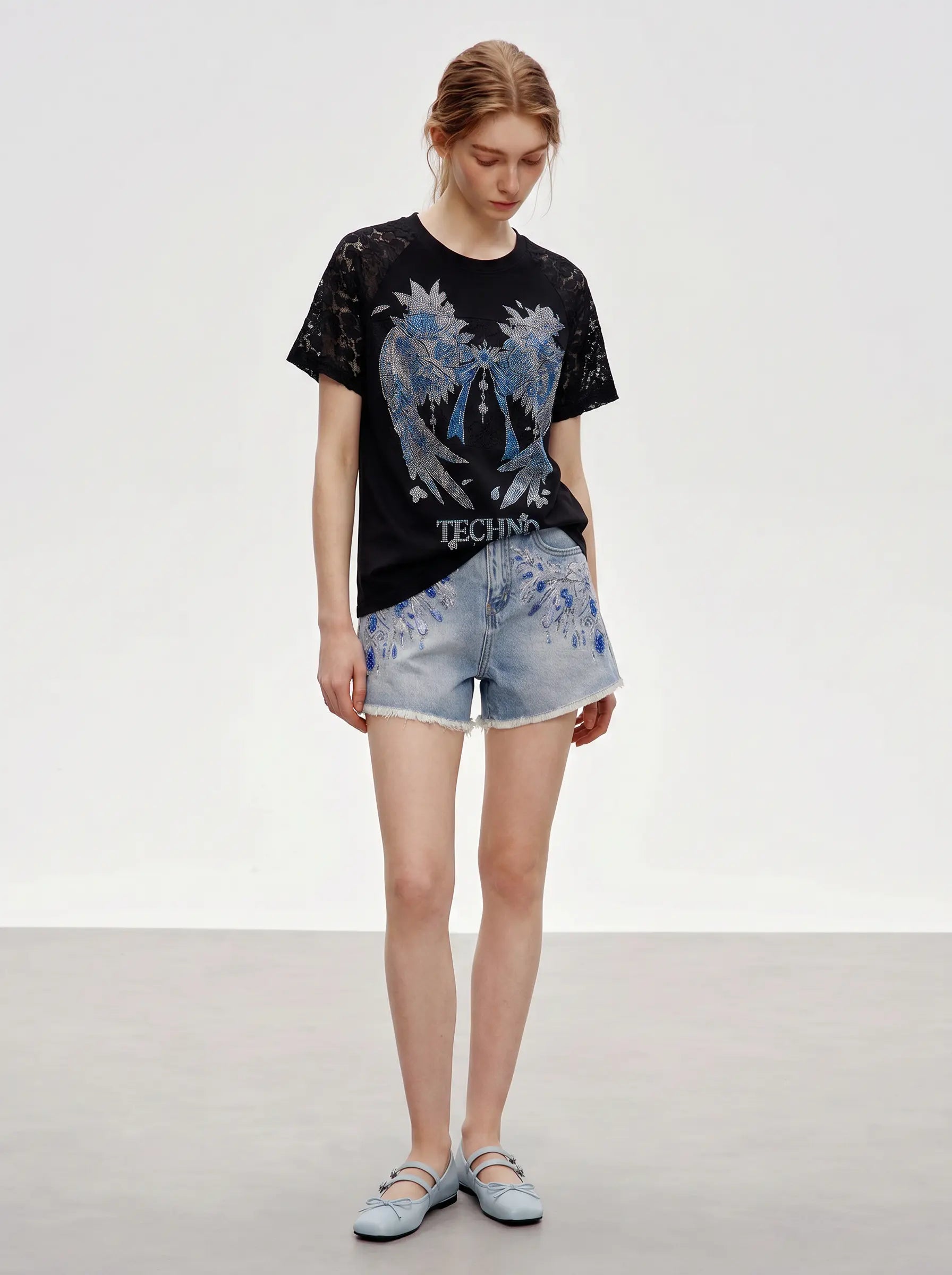 A model stands against a plain light gray studio background wearing a black graphic T-shirt with a large blue angel design and the word 'TECHNO' on the front, paired with light blue denim shorts and pale blue flats. TCH Rhinestone Floral Wing Techno Graphic T-Shirt